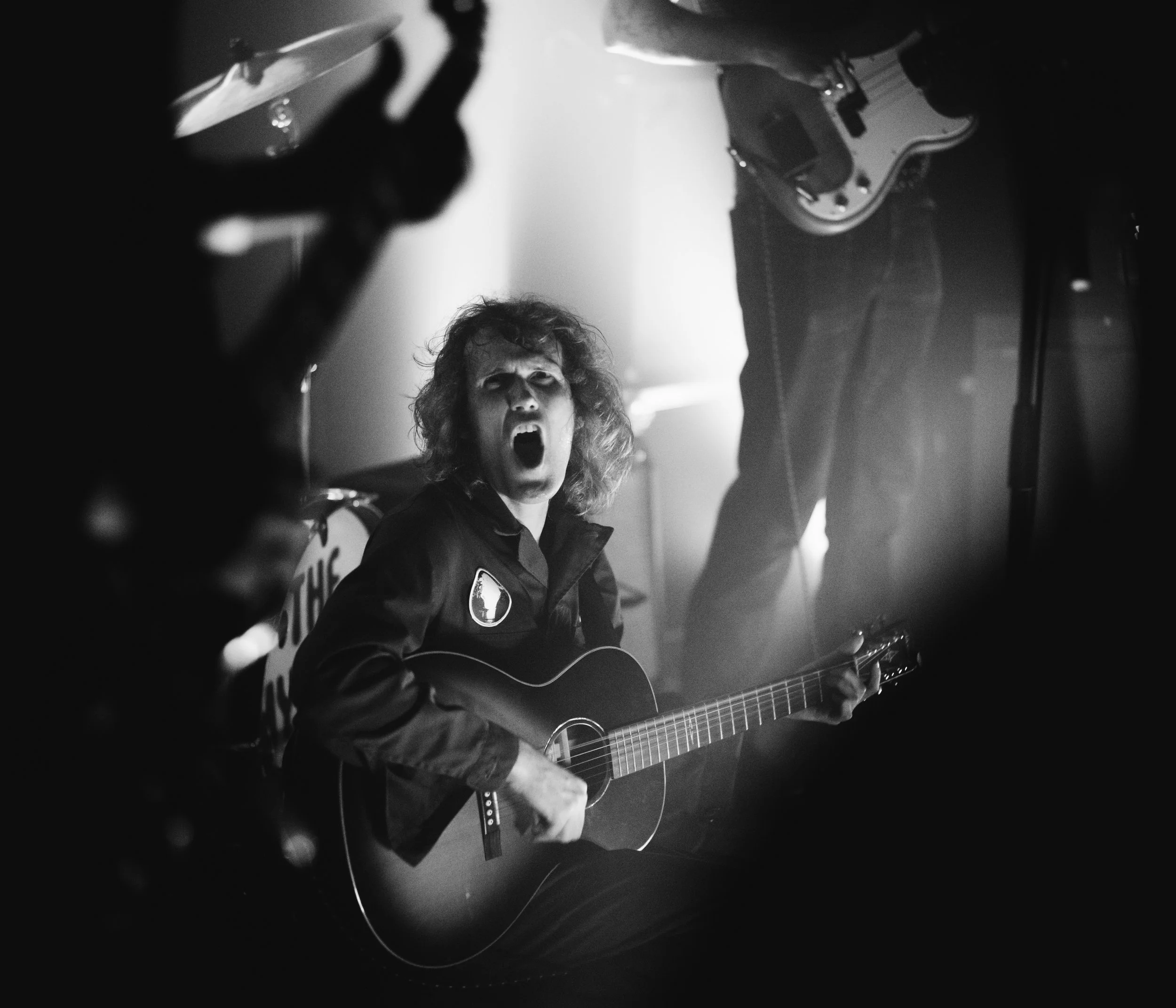 A black and white photo of a person with curly hair and a serious expression, singing and playing an acoustic guitar in a dimly lit setting. The person appears to be immersed in music, with a background featuring musical equipment and instruments.