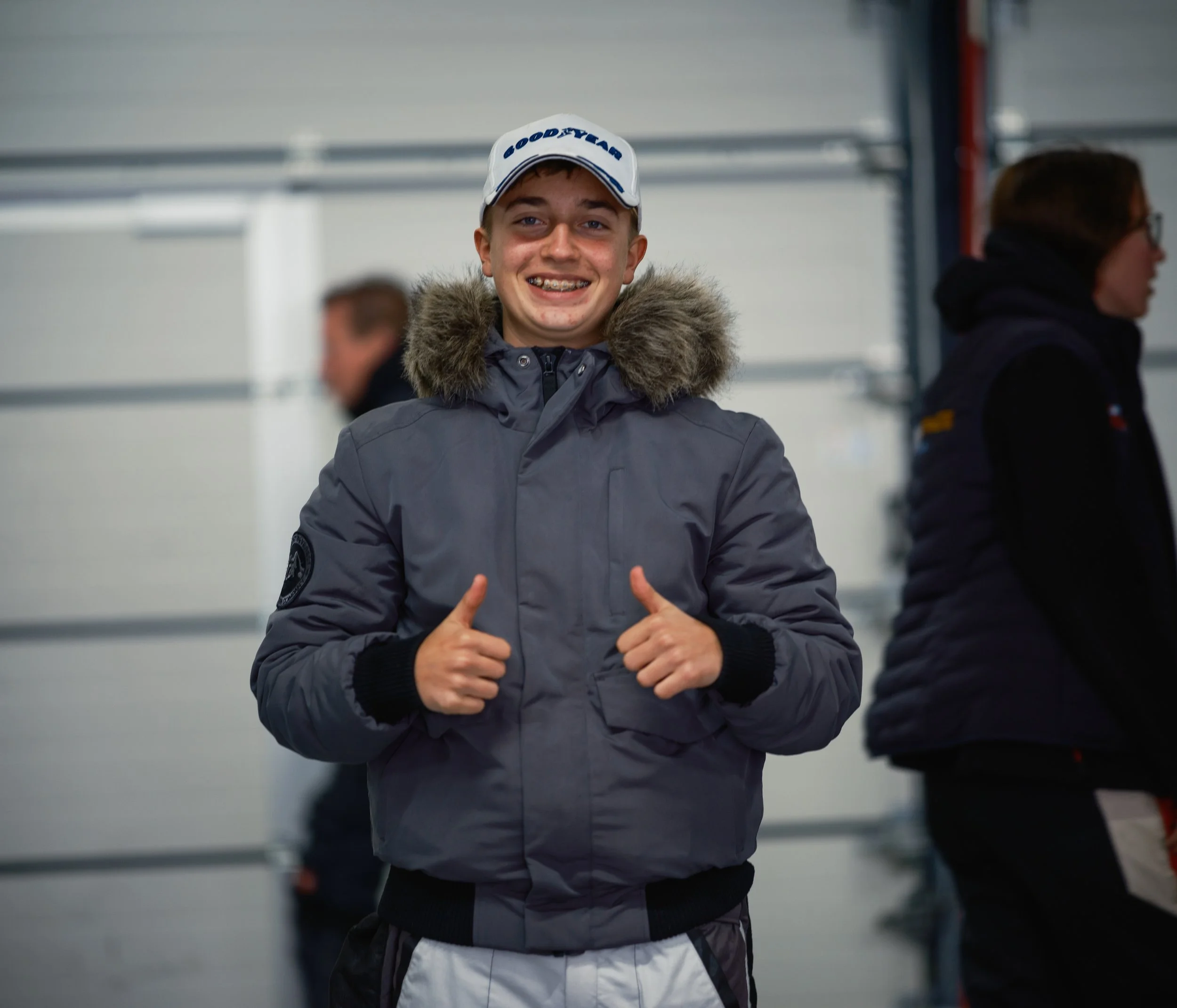 Smiling young man with braces wearing a gray winter jacket and a white Goodyear cap, giving two thumbs up in an indoor setting with blurred people in the background.