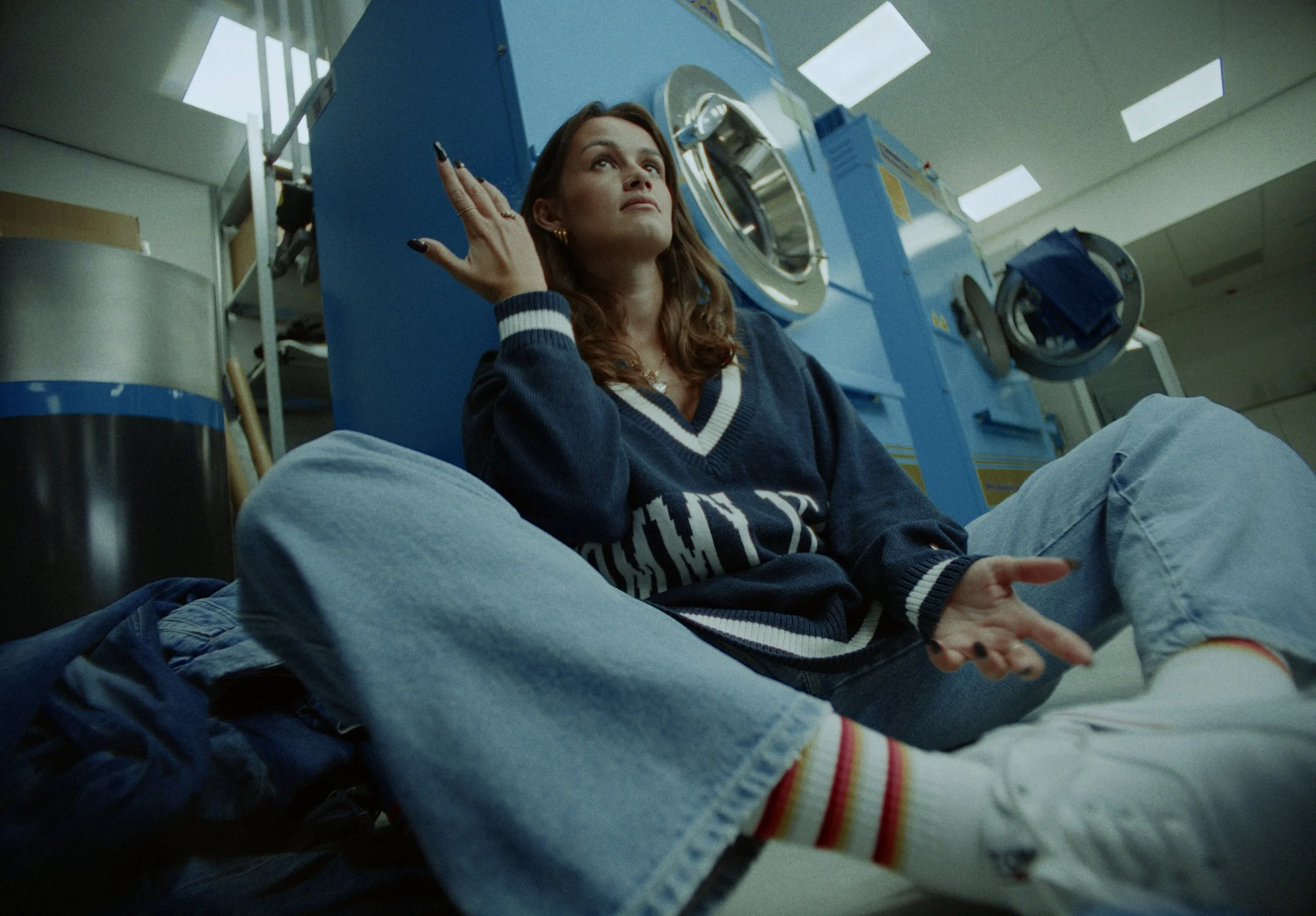 A woman sitting on the floor in front of a large washing machine in a laundromat, looking upward with her hand touching her head.