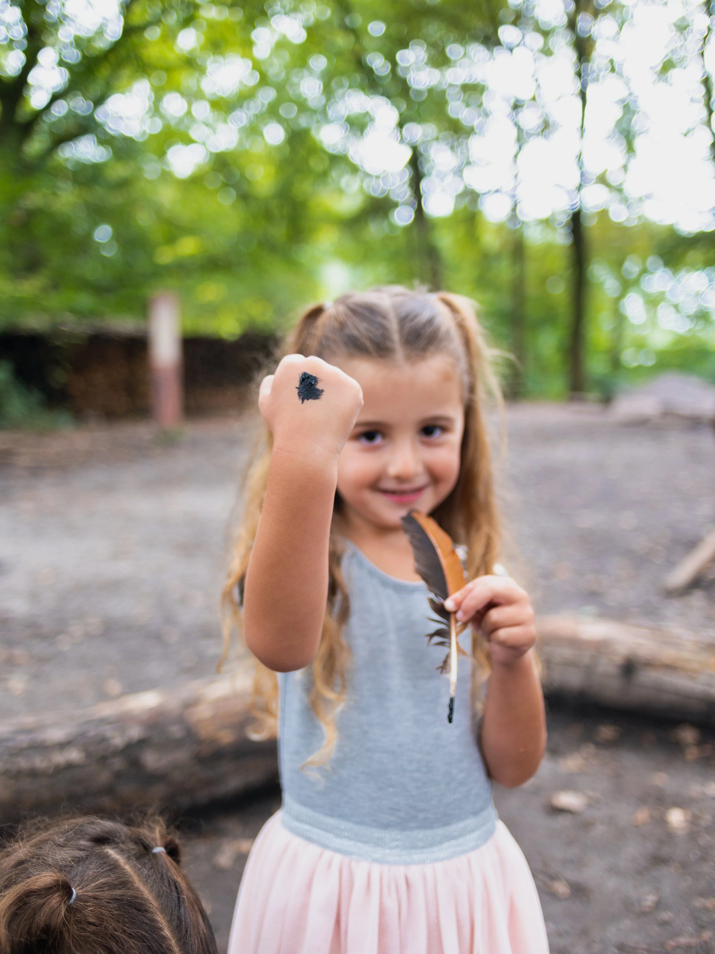A young girl with long hair in pigtails holding a feather and a piece of charcoal, smiling and showing her fist with a black makeup mark, outdoors with trees and blurred background.