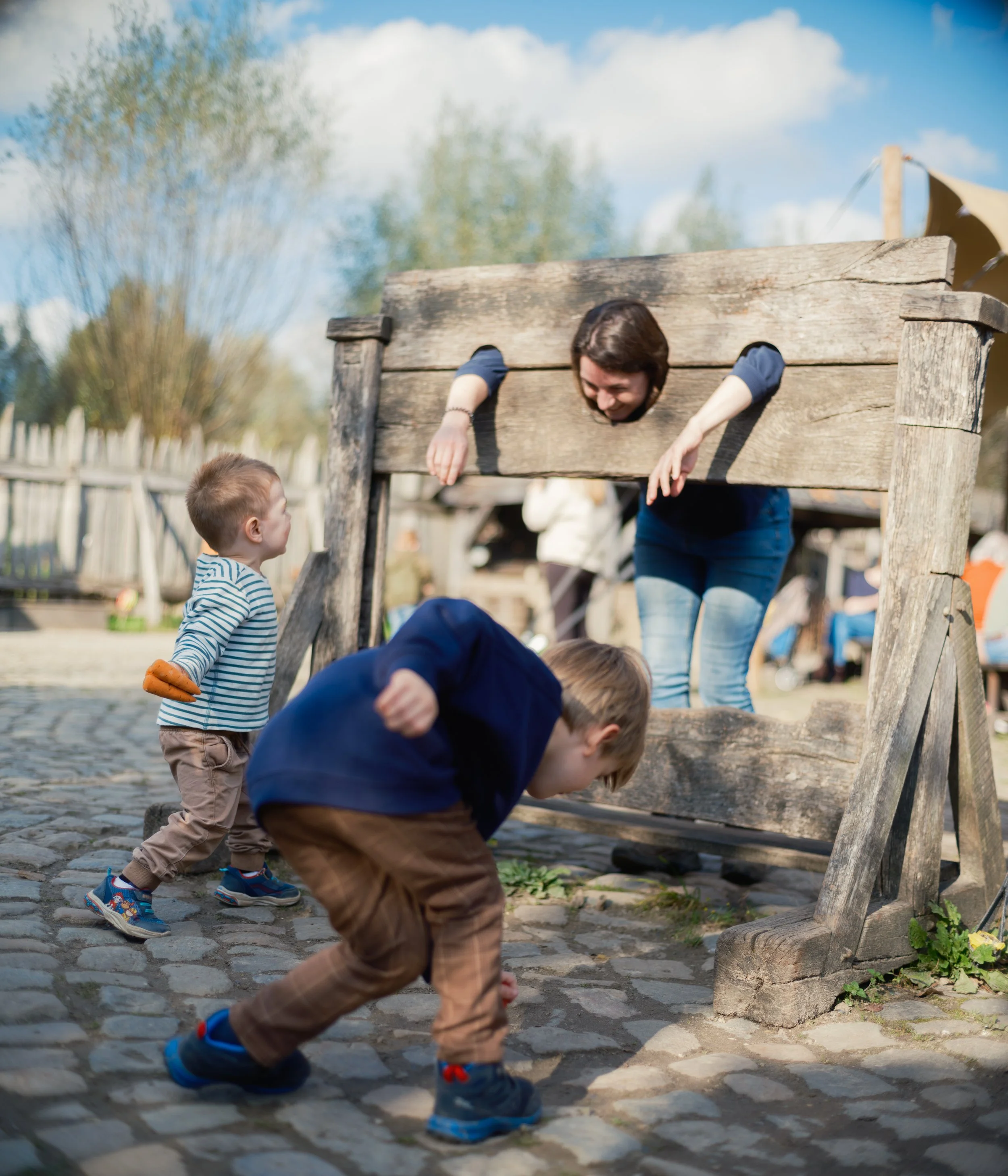 Children playing outdoors, with one girl climbing through an old wooden structure and a boy crouching nearby, on a cobblestone surface under a partly cloudy sky.