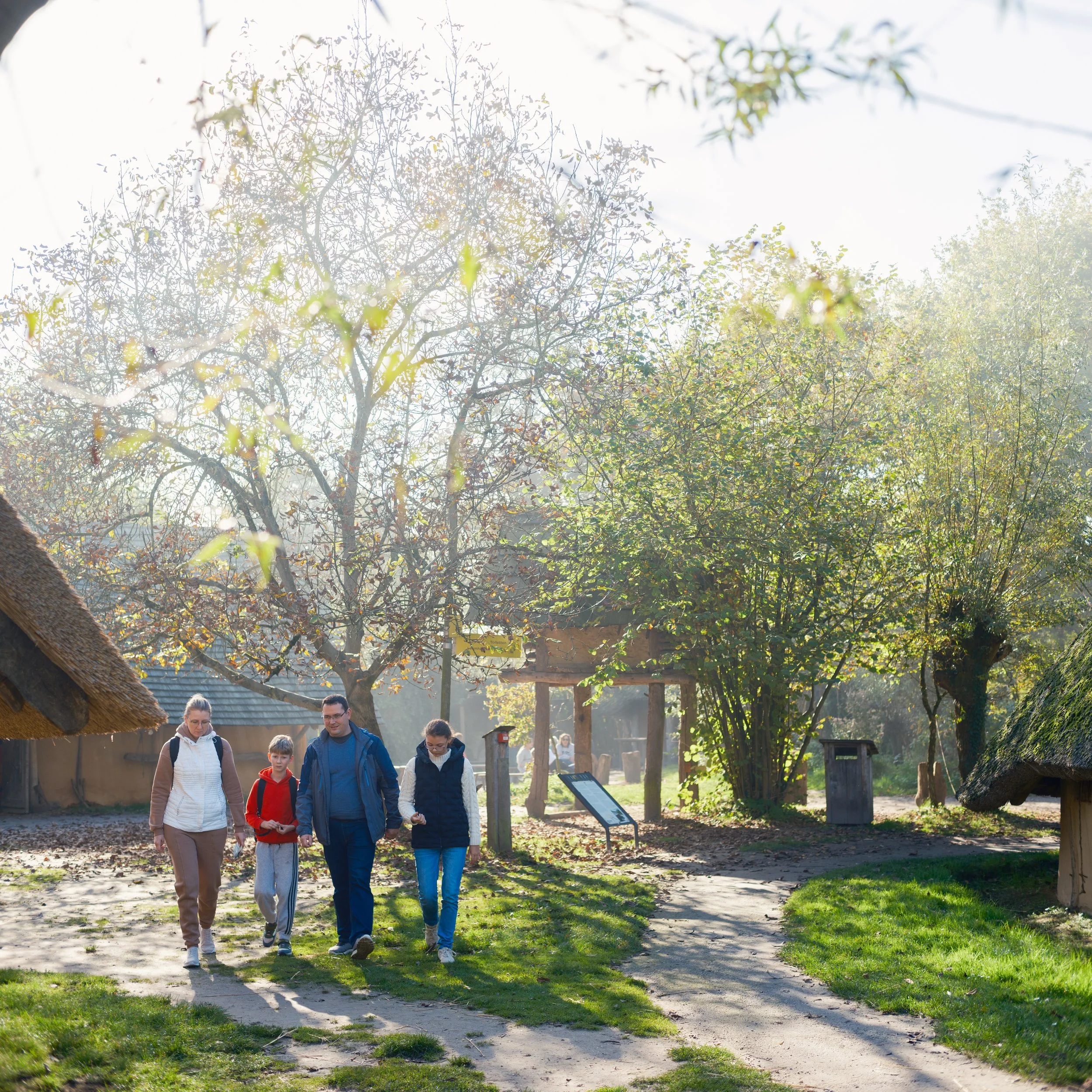 A group of people walking outdoors in a park or garden on a sunny day, with trees and a thatched roof structure nearby.