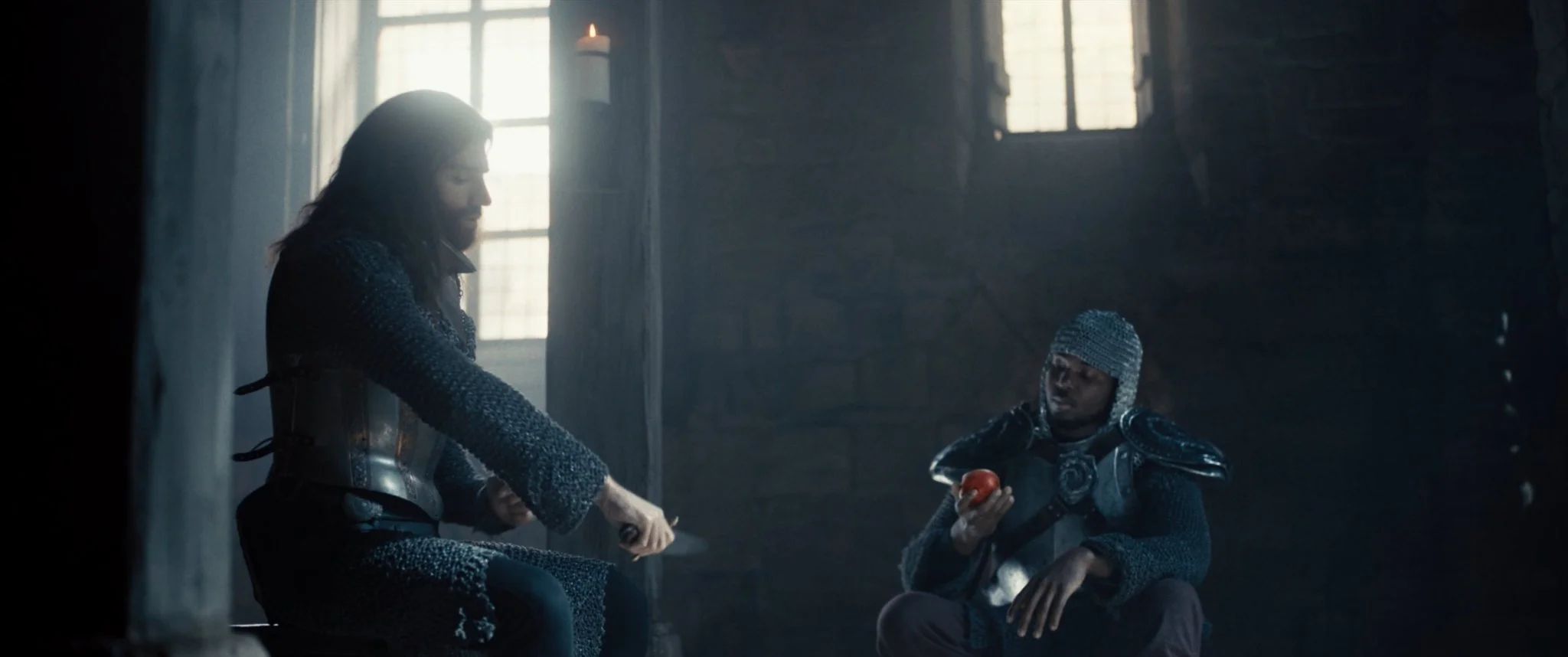 Two medieval characters in chainmail armor sit in a dimly lit stone room with large windows. One character, with long hair, is holding a sword, while the other, wearing a helmet, is holding a tomato.