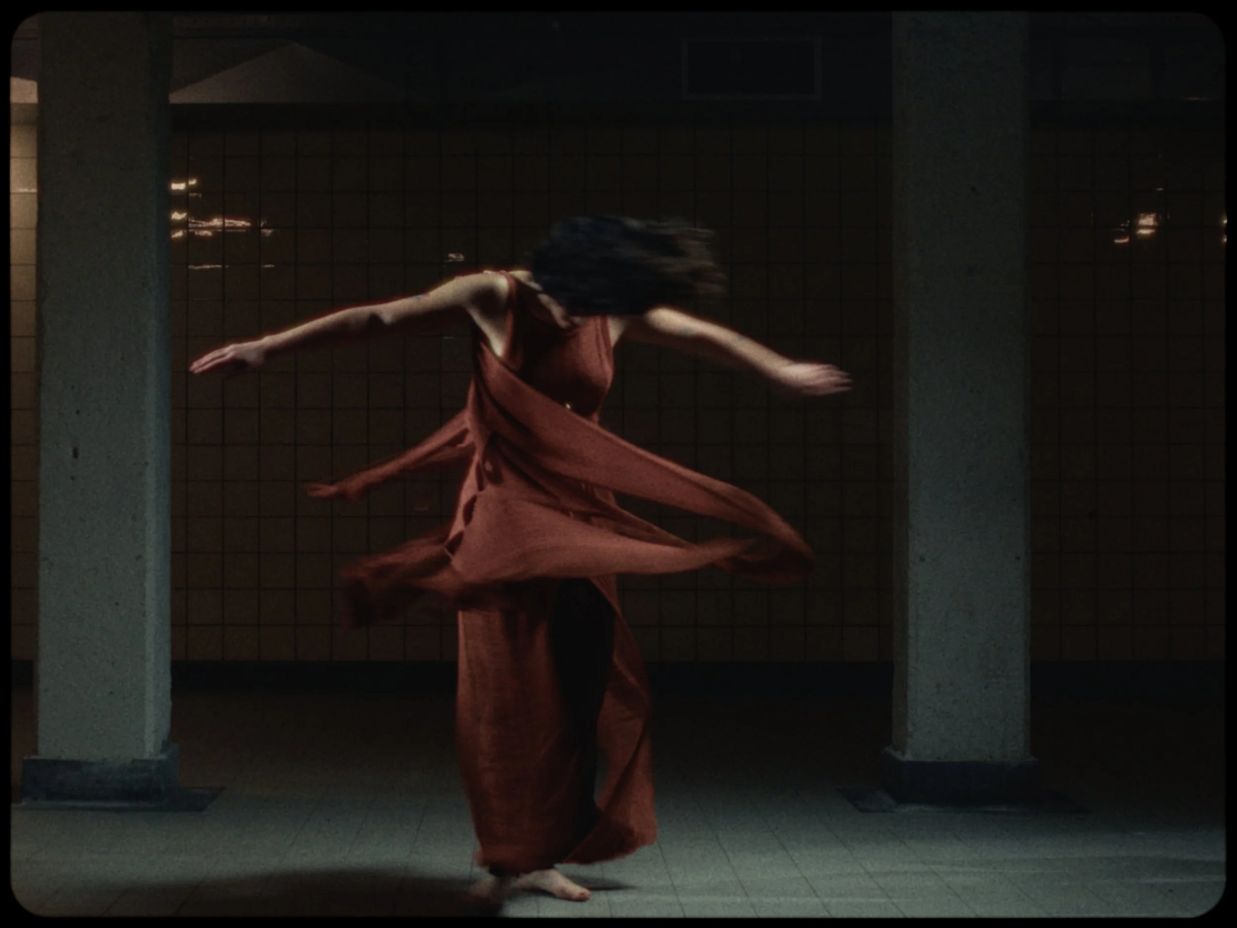 A woman dancing in a long, flowing orange dress in an indoor space with tiled floors and concrete pillars.