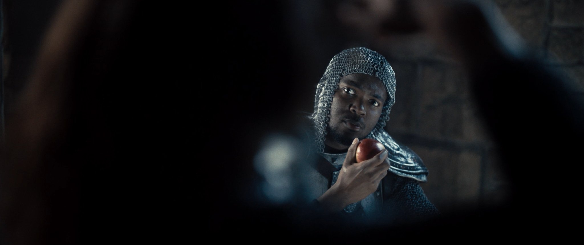 A man dressed as a knight with a chainmail hood looking through a hole, holding a red apple.