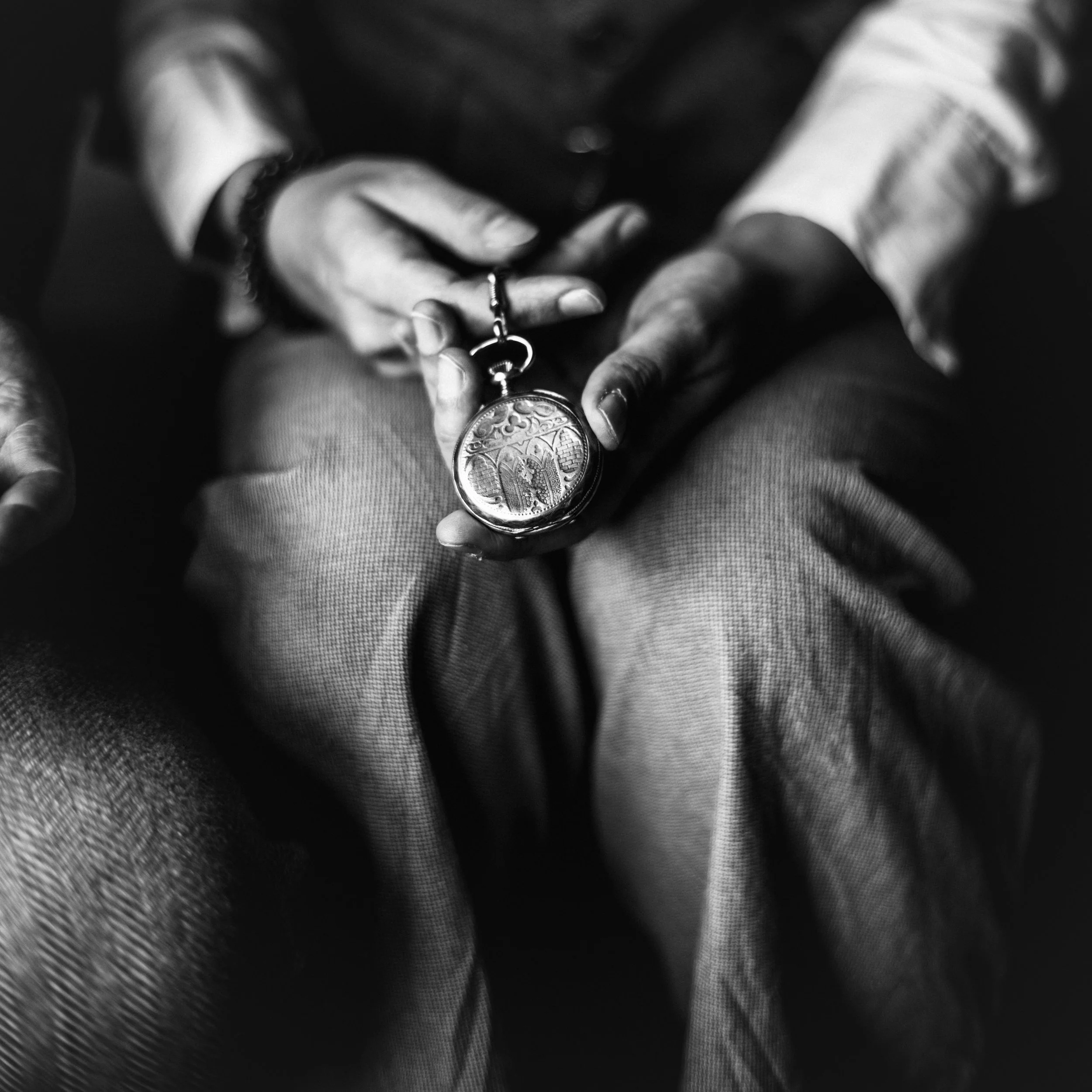 Close-up of a person's hand holding a pocket watch, focused on the intricate details of the watch, with a blurred background and person's lap, in black and white.