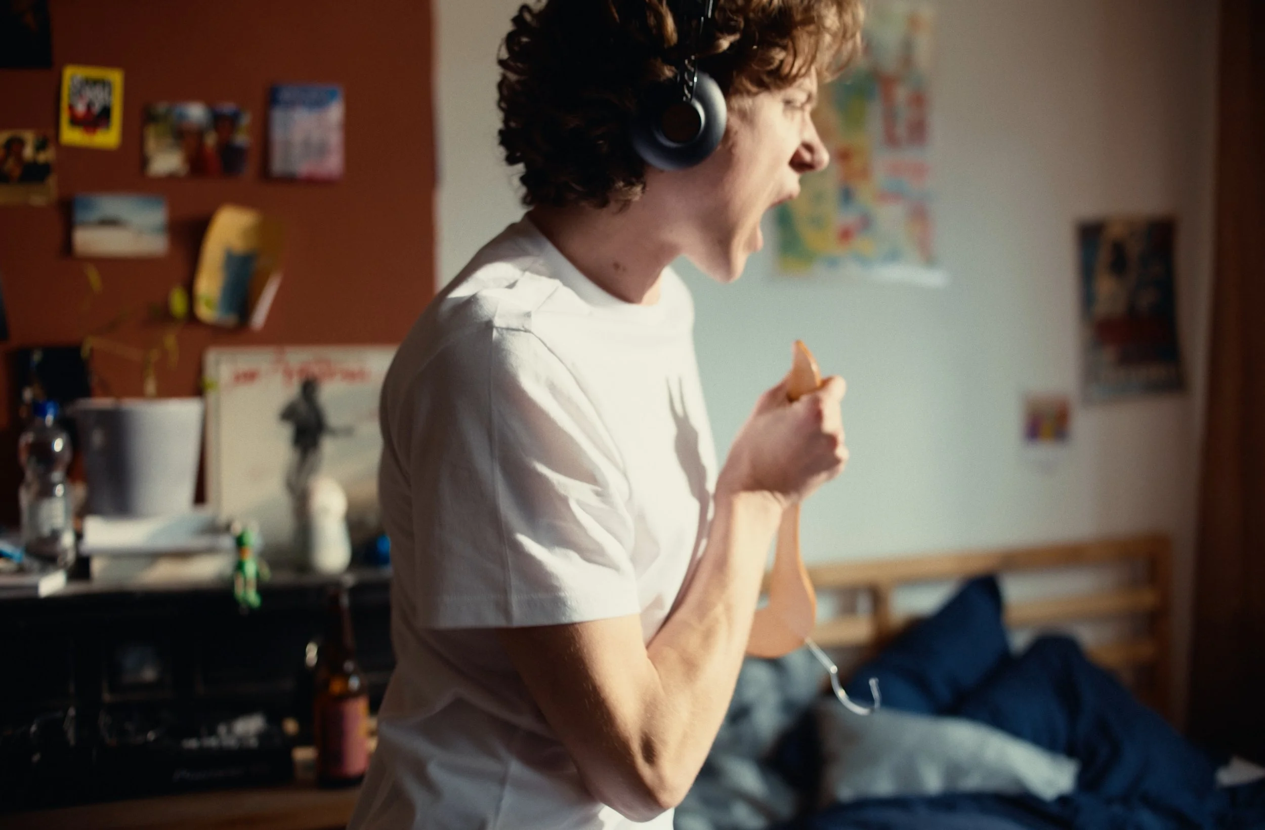A person with curly hair yelling or singing passionately while wearing headphones and holding a stick or chopstick in a room with various pictures and memorabilia on the wall.