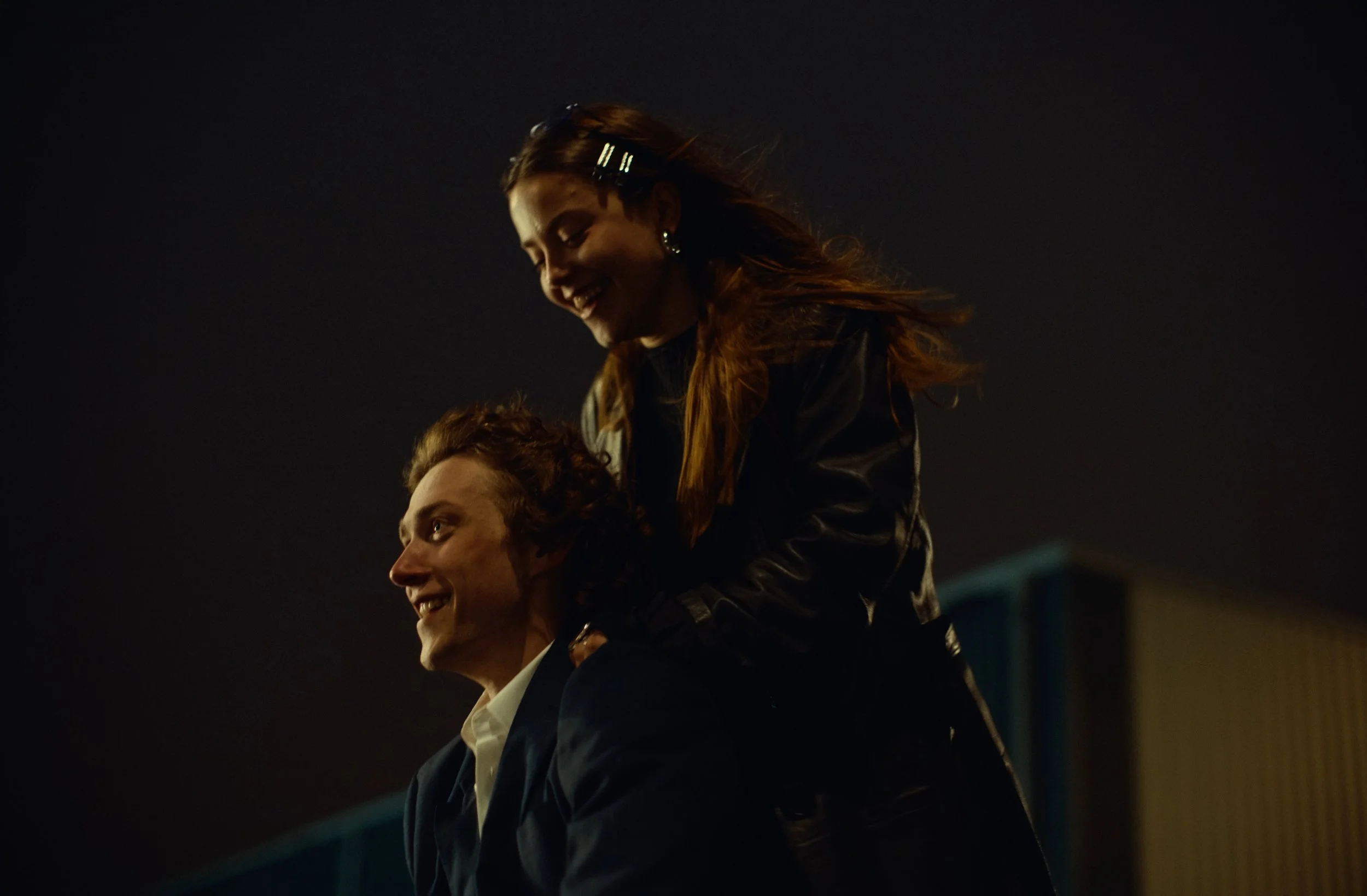 A woman with long hair and earrings riding on the shoulders of a man in a suit during nighttime. They are outdoors under a dark sky.