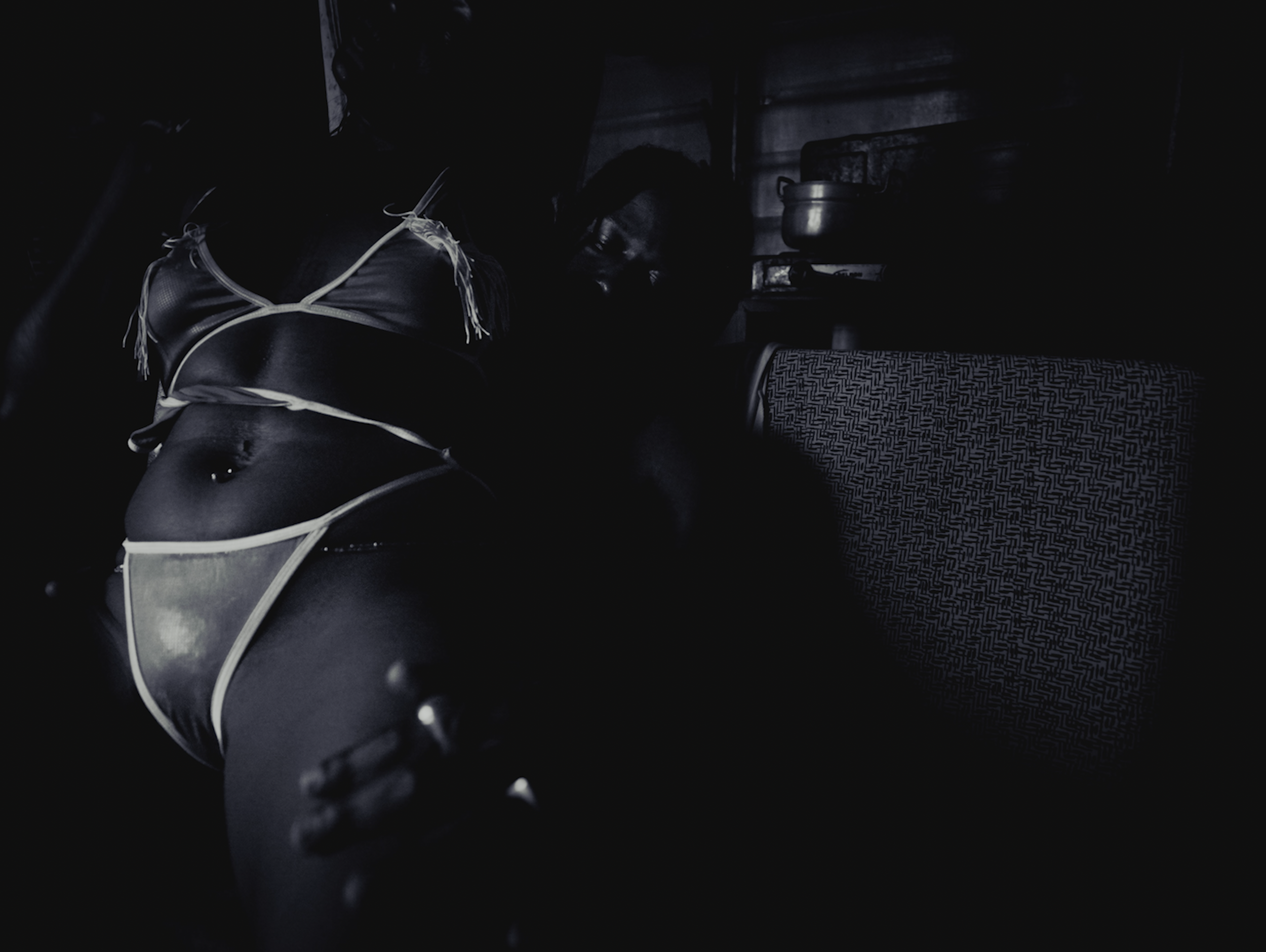 A person lying down in a dark room, wearing a black and white outfit, with tattoos visible on their body, near a table with patterned fabric and some kitchen pots.