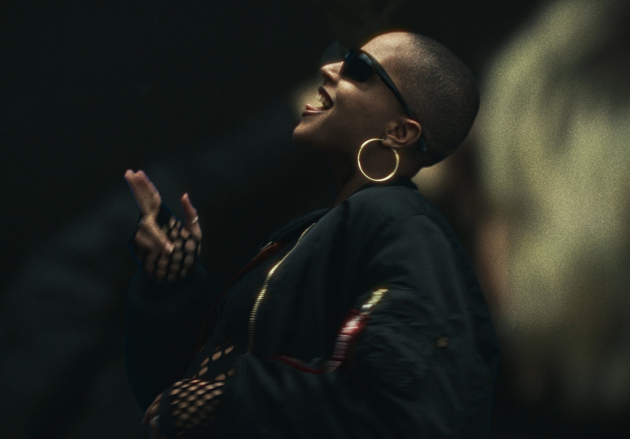 A woman with shaved hair and wearing sunglasses, hoop earrings, a black jacket, and fishnet gloves, is smiling and displaying her middle finger.