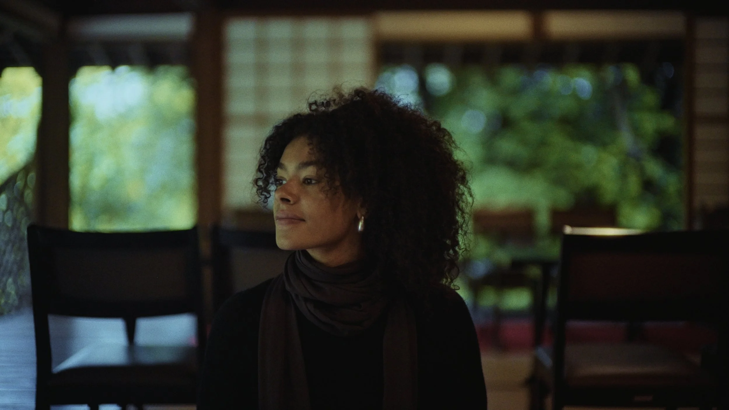 A woman with curly hair and hoop earrings in a dark top and brown scarf, looking to her left, sitting inside near large windows with green foliage outside.