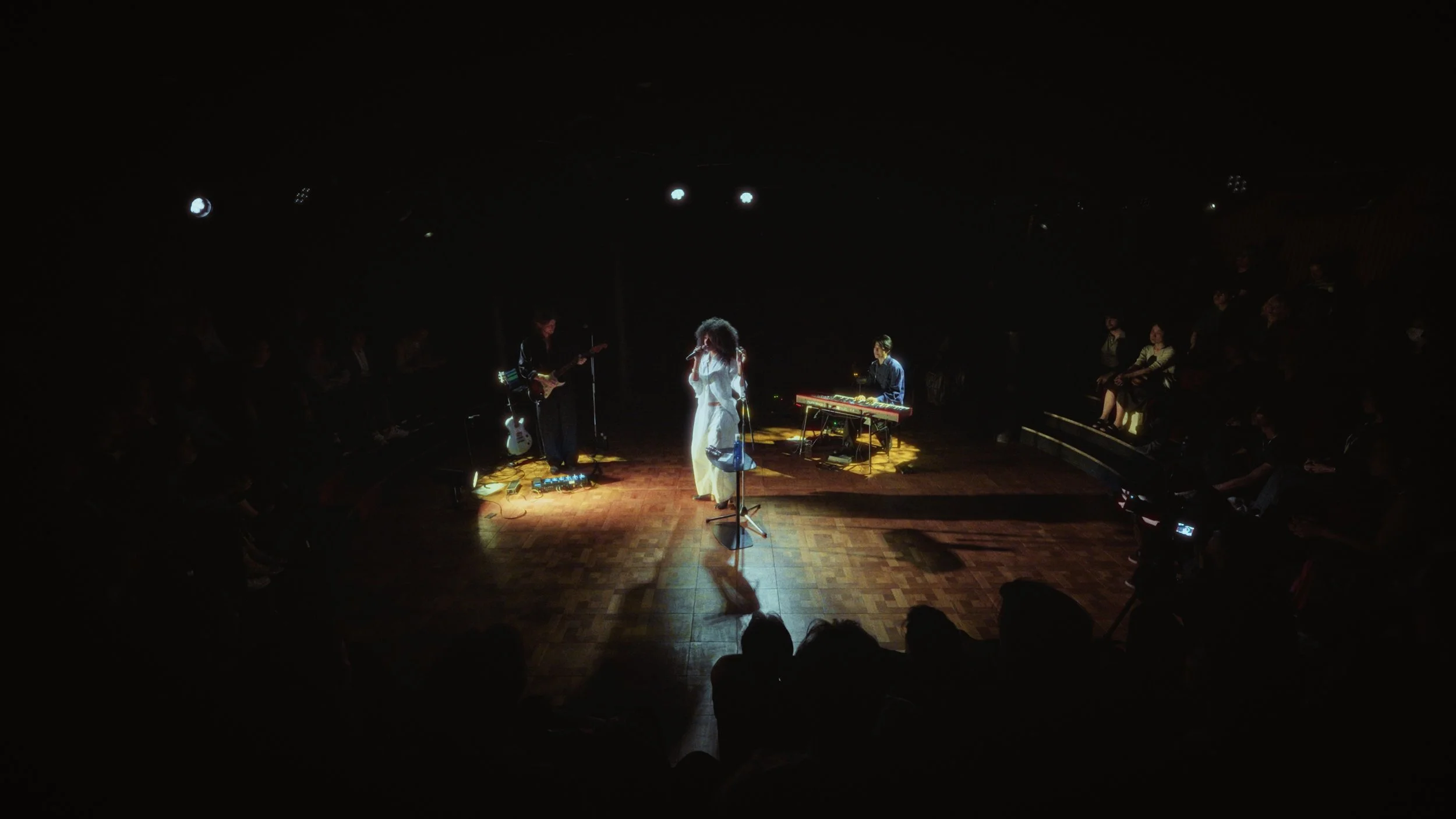 A musical performance on stage with a singer, guitarist, keyboardist, and audience watching in a dark venue.