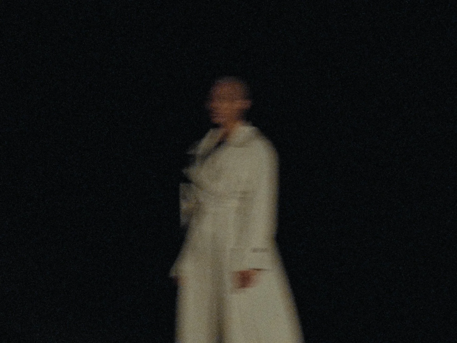 A person standing in a dark environment, wearing a light-colored jacket or coat, with short hair, smiling or laughing.