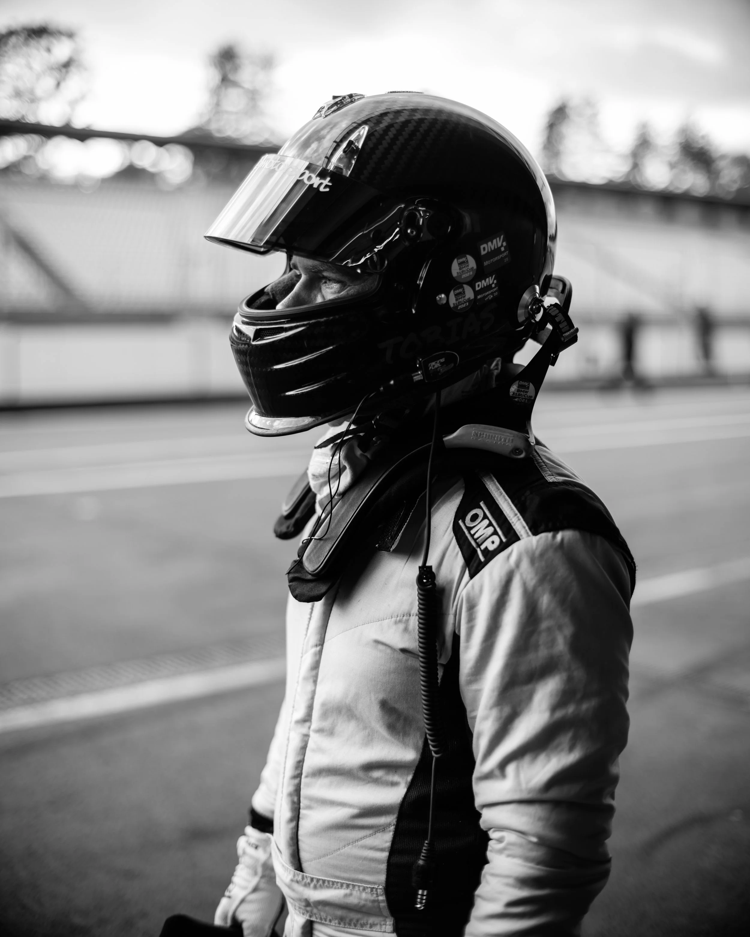 A race car driver wearing a racing helmet and racing suit, standing on a race track.