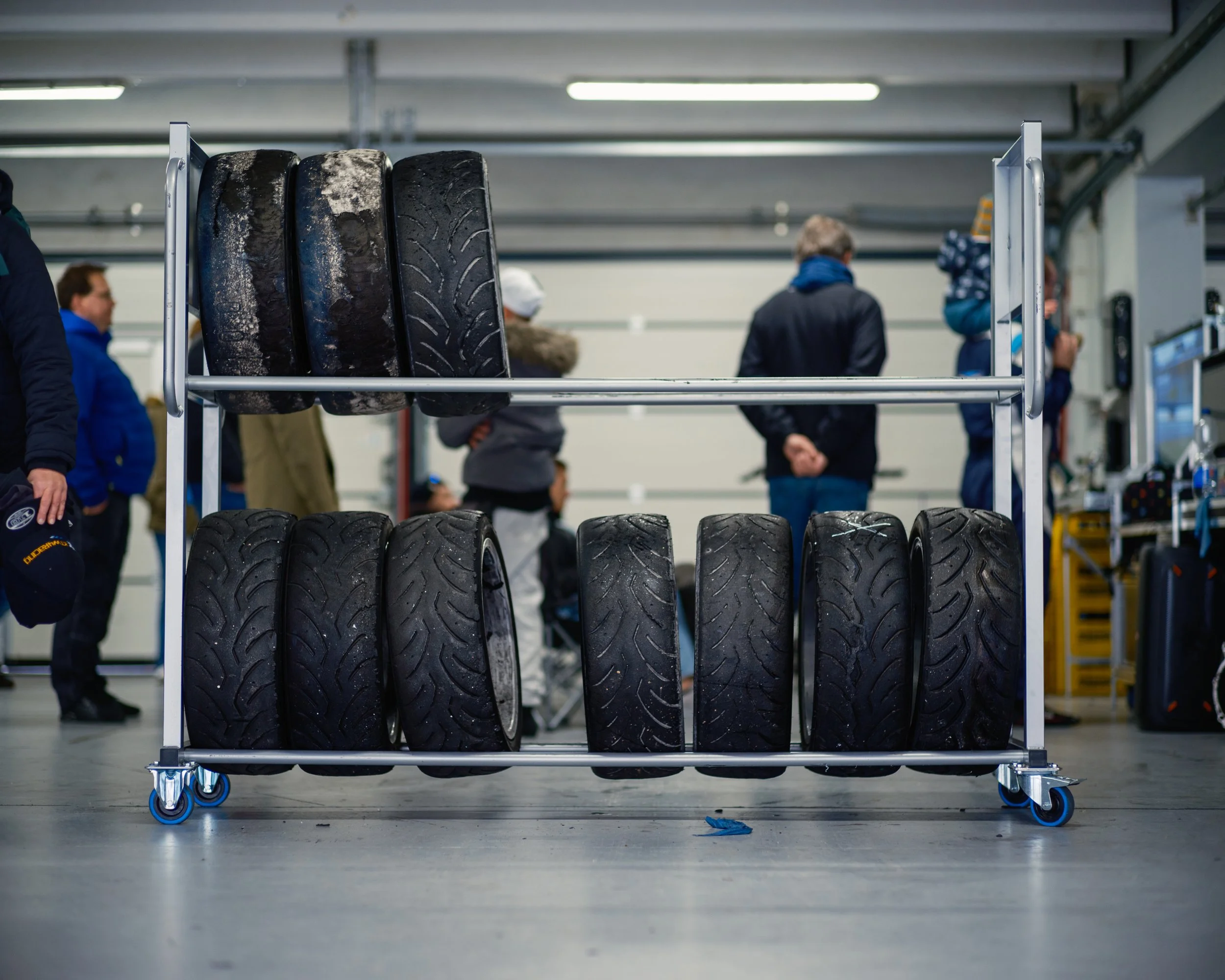 A metal rack with four worn tires on a blue wheeled cart in a garage or warehouse, with people in the background