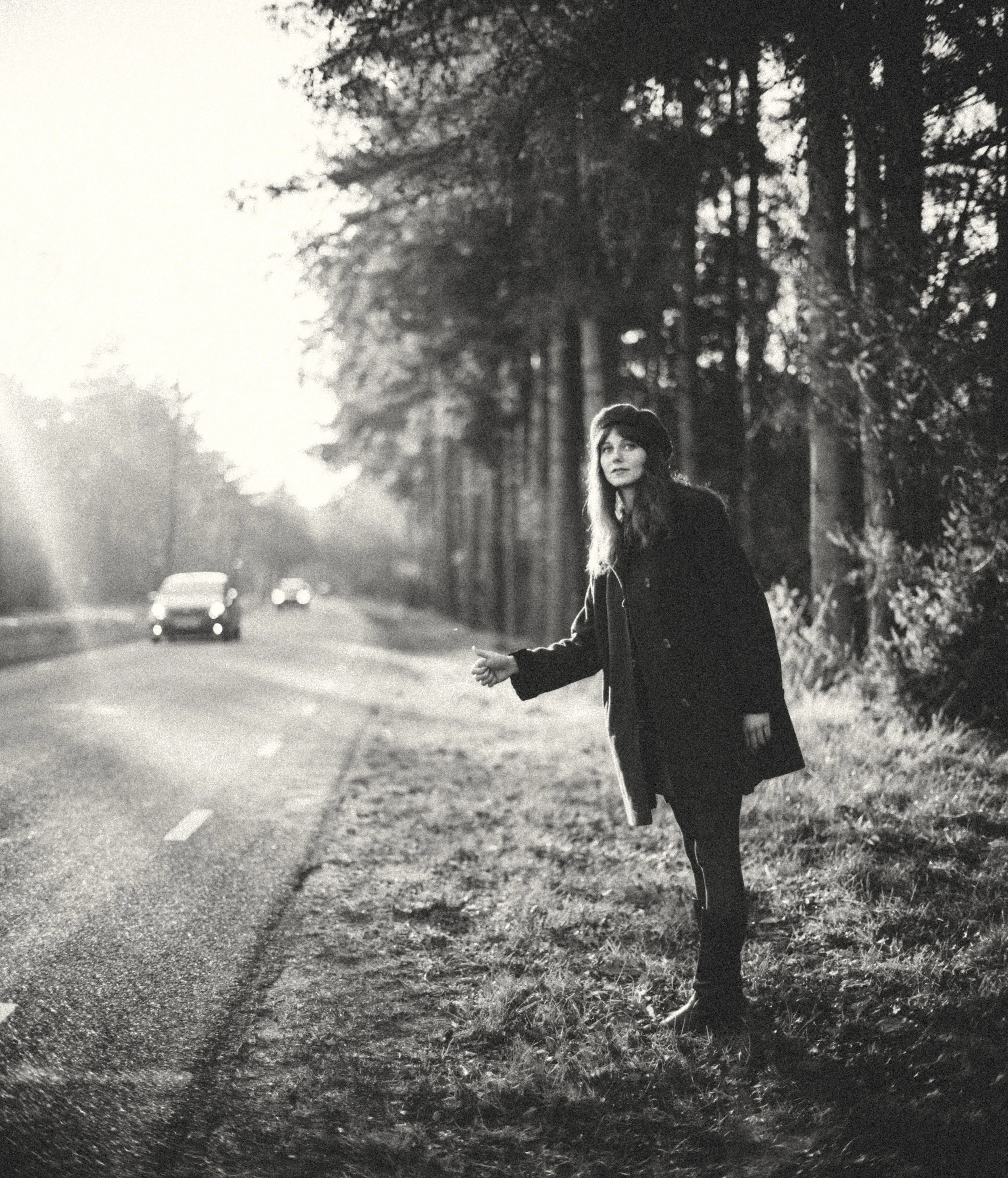 A black and white photo of a woman standing next to a tree on the side of a road, holding a book in her hand, with cars driving in the background.