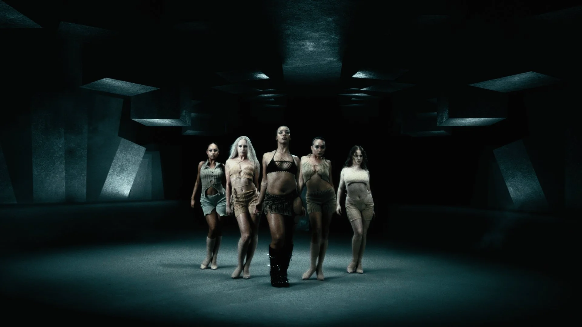 Five women walk confidently in a dark, industrial-looking space with geometric ceiling structures, wearing revealing, stylish outfits.