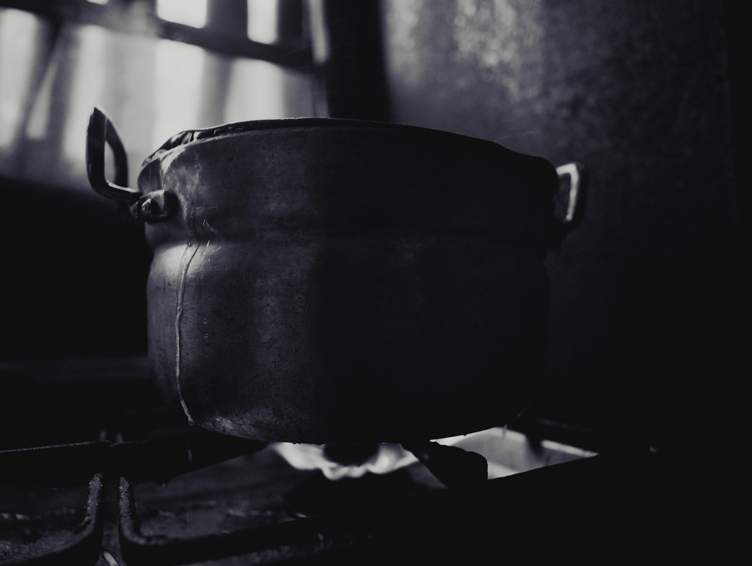 A metallic kettle on a stovetop with a dark surface, wooden handle and spout, under a window with sunlight streaming through.