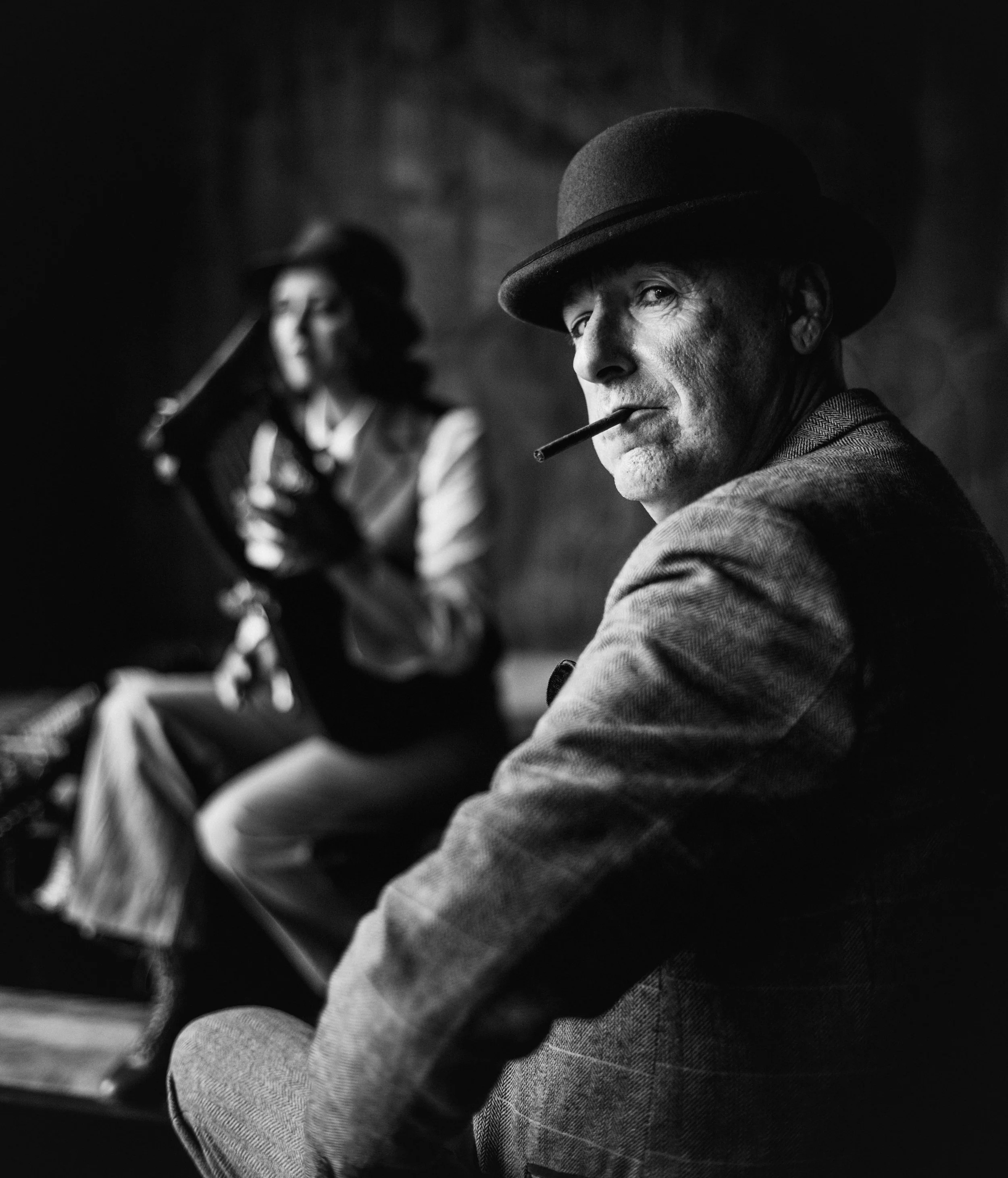 A black and white photo of a man with a fedora hat smoking a cigar, with a woman in a hat out of focus in the background.