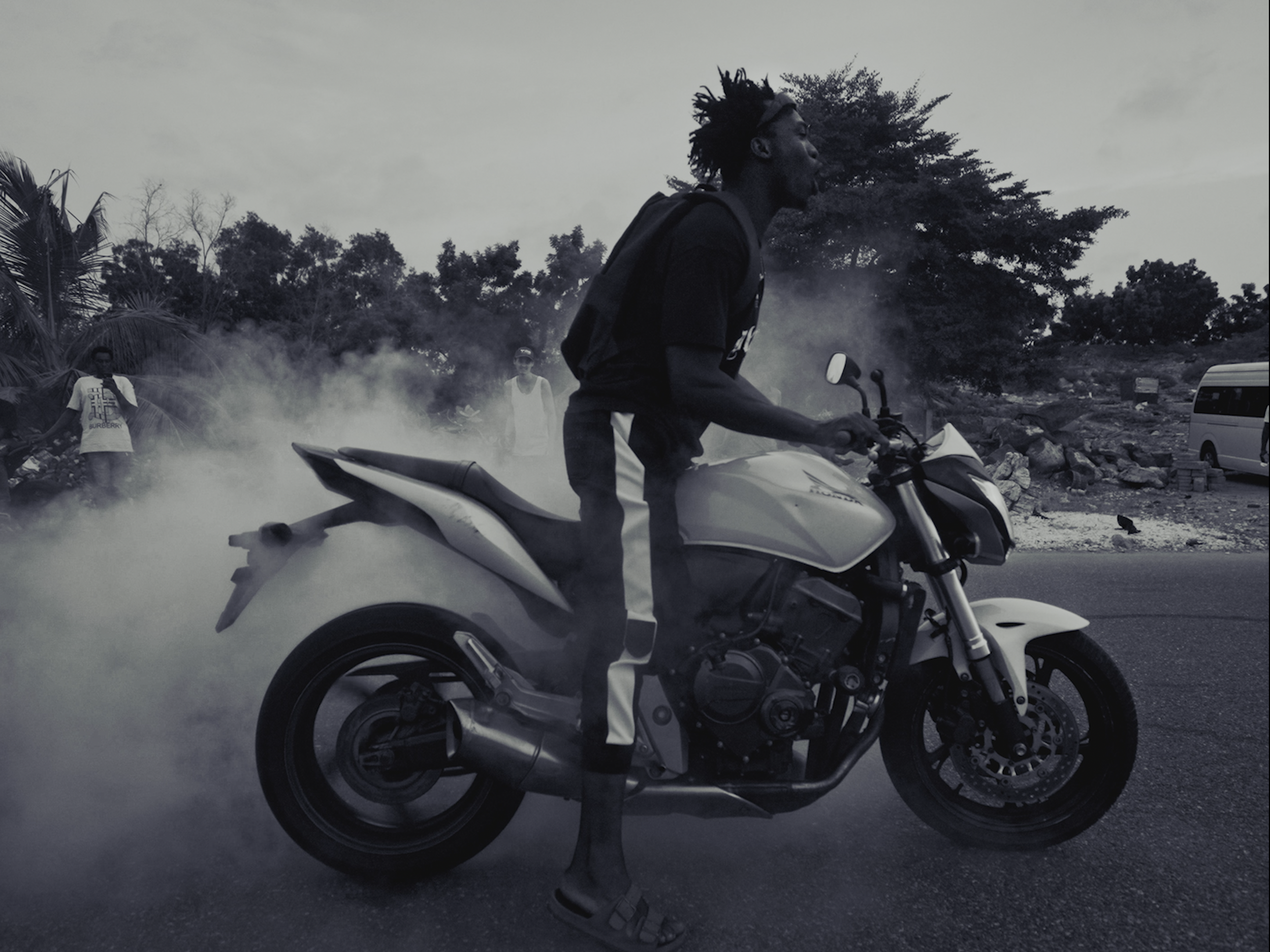 A man with dreadlocks riding a motorcycle, surrounded by dust and trees in the background.