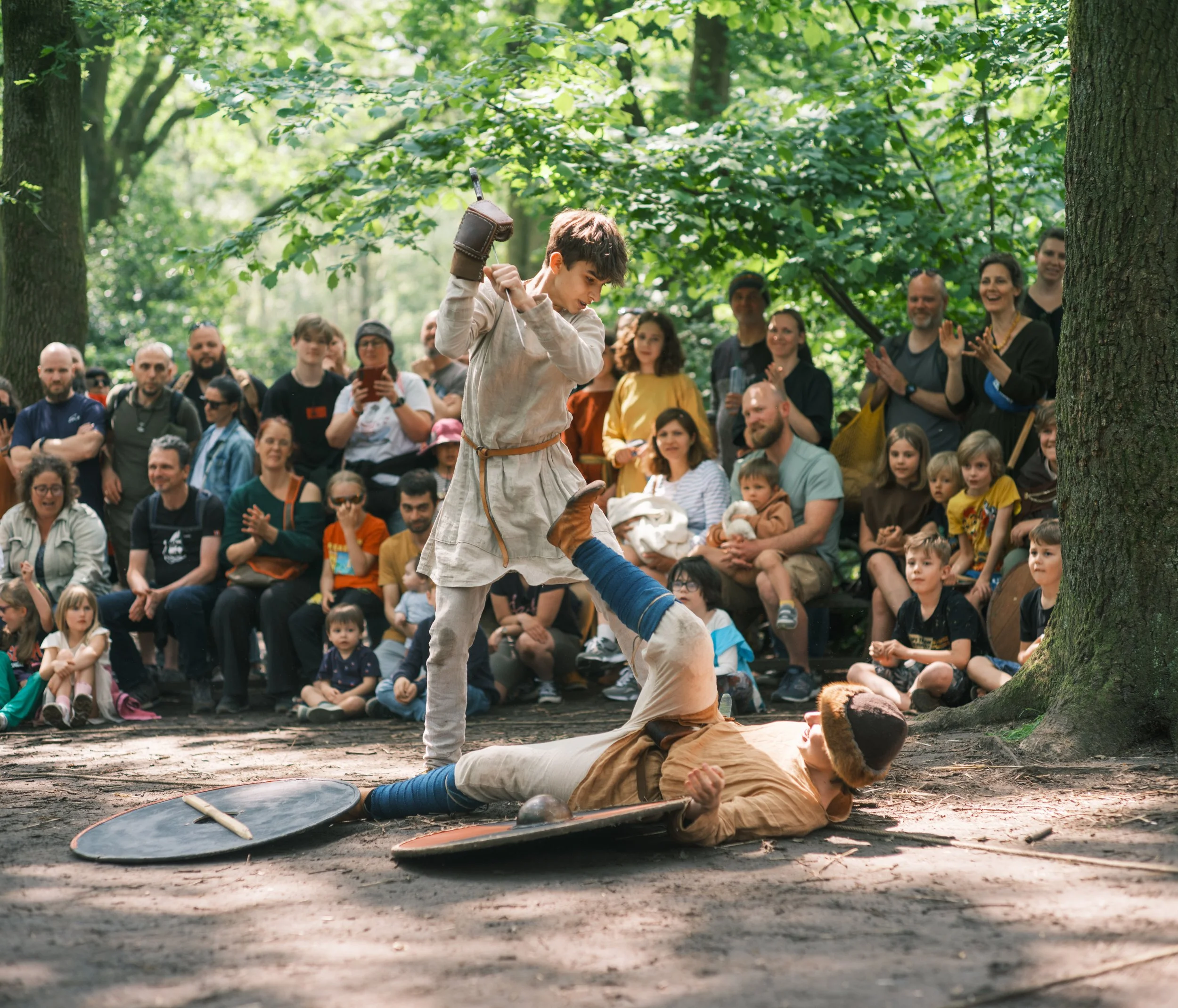 A reenactment of a sword fight in a wooded area with a crowd watching. A young boy dressed in medieval costume is jumping on a person lying on the ground, engaging in the activity with a sword.