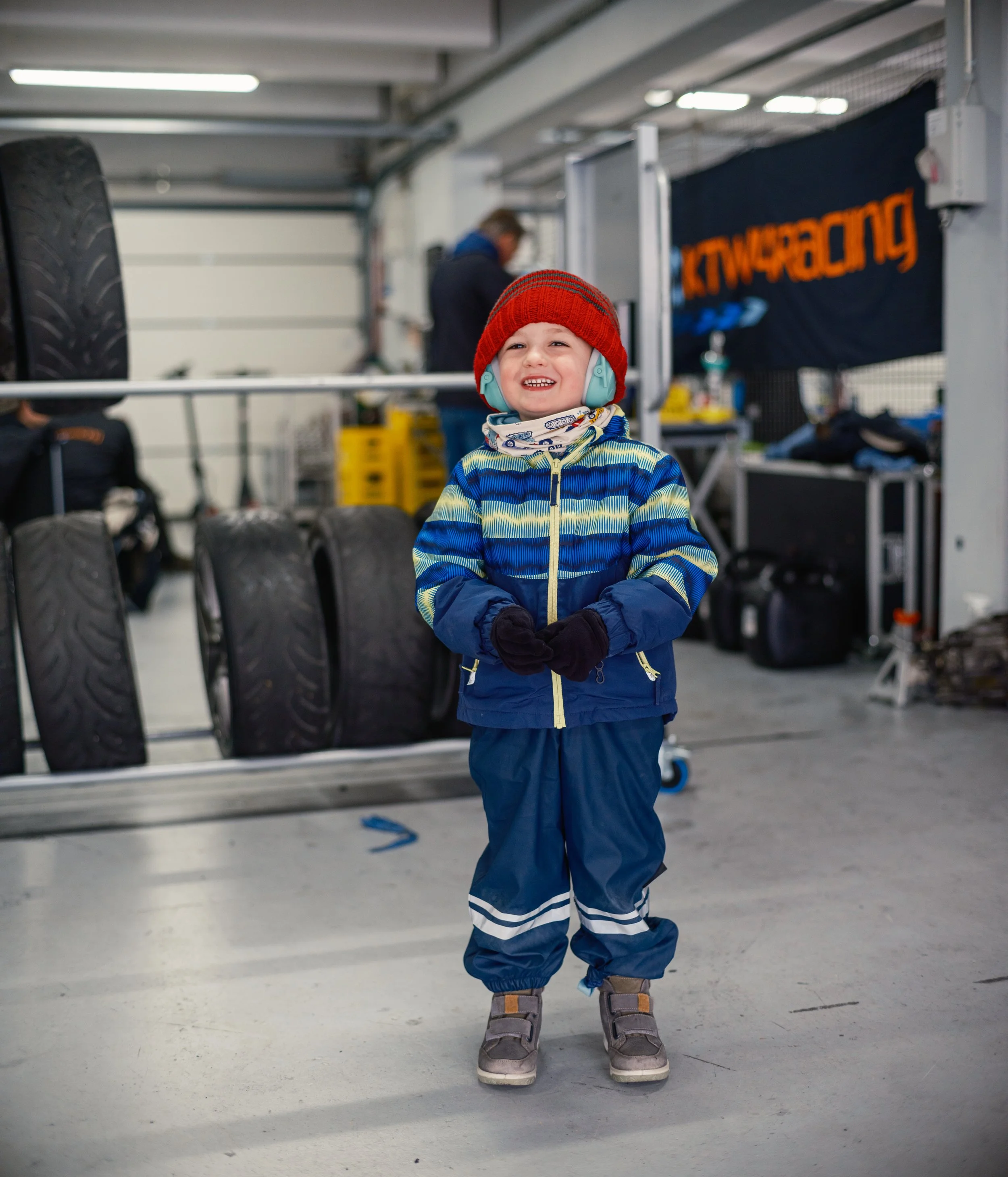 A young boy dressed warmly in an oversized blue and yellow striped jacket, blue snow pants, gray boots, a red knit hat, and light blue headphones, smiling in a garage or workshop area with tires and tools in the background.