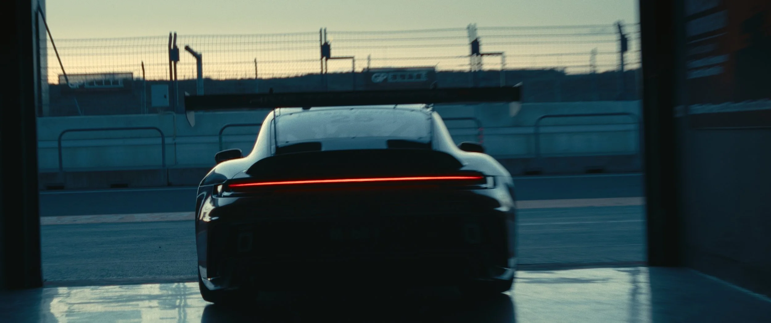 A black sports car with a large rear wing, seen from behind, on a race track during sunset.