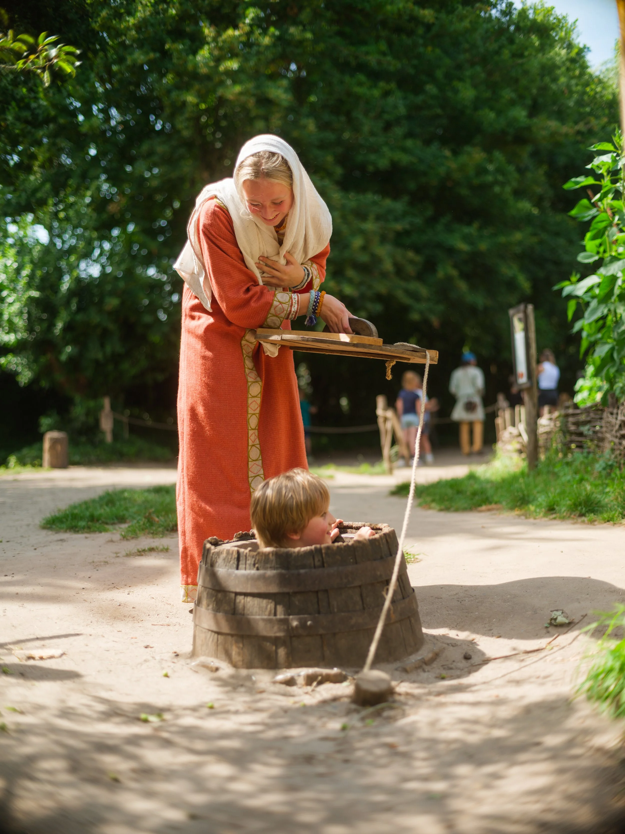 A woman in a long orange dress with decorative patterns, covered with a white scarf, smiles while pushing a boy in a wooden barrel with a string. The boy is holding onto the string, and both are outdoors in a park or garden with lush green trees and 