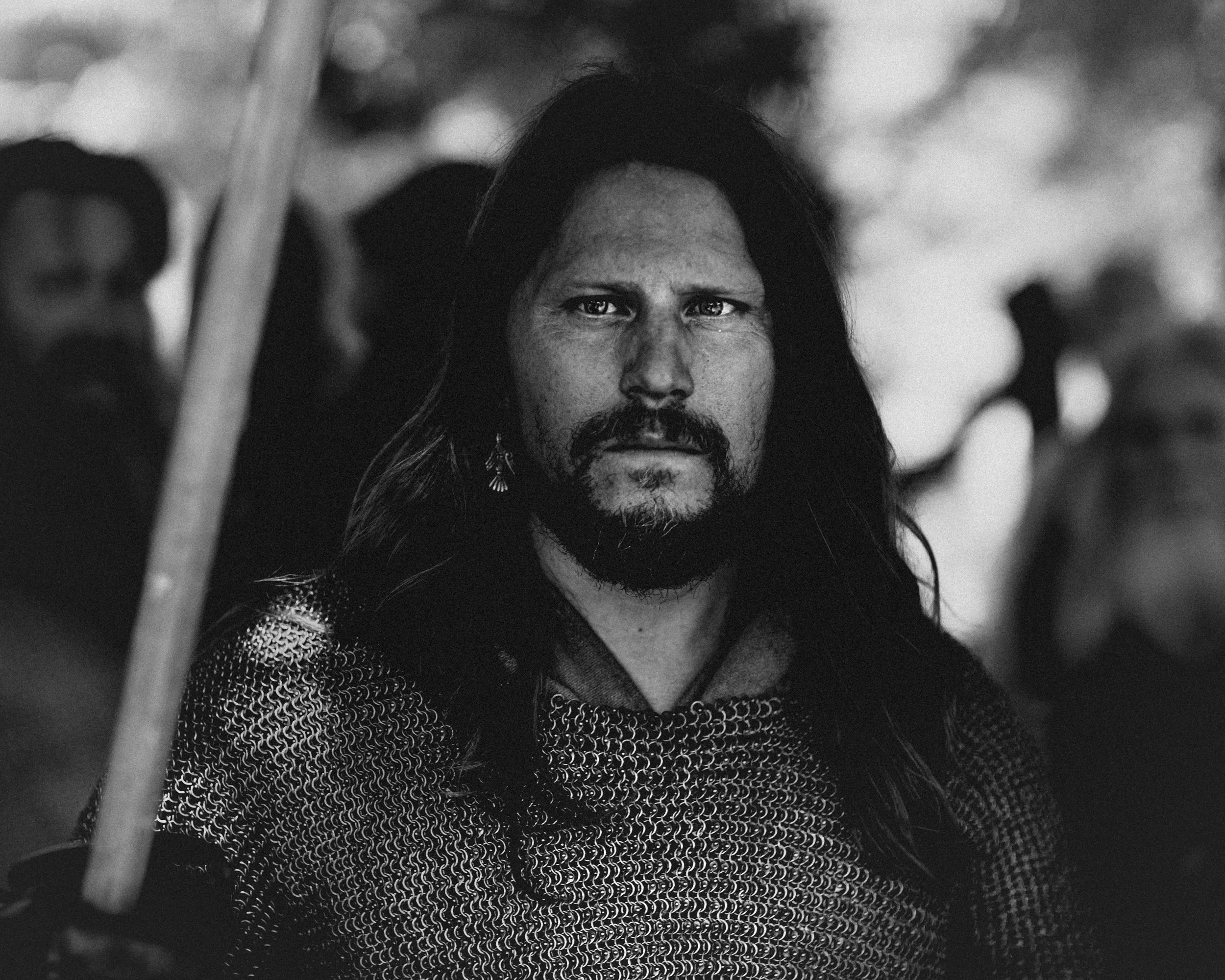 A person with long dark hair, a beard, and earrings wearing chainmail armor, facing forward with a serious expression, in a black-and-white photo.