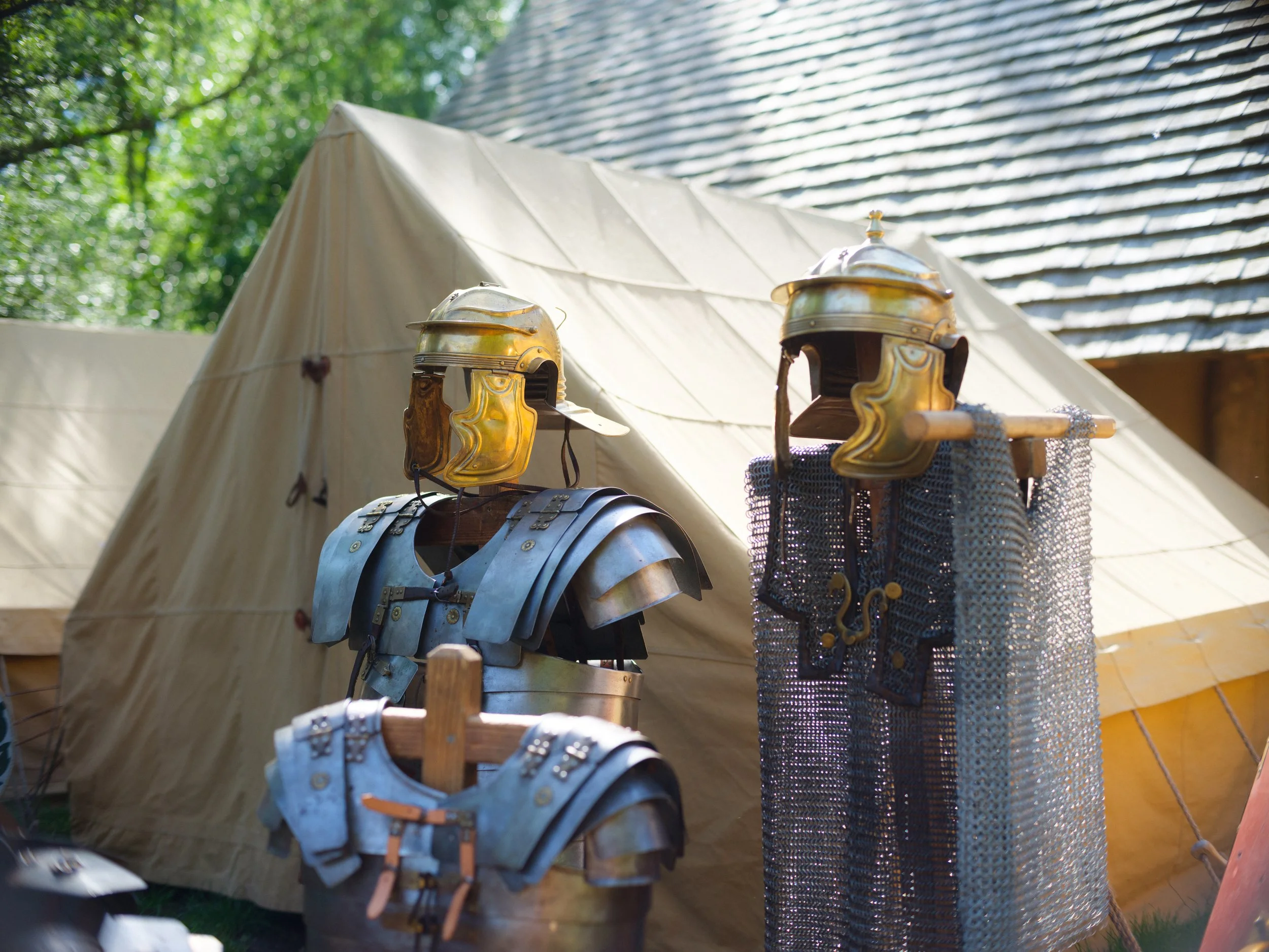 A beige canvas tent with medieval armor and helmets displayed outside on a wooden rack, with green trees and sunlight in the background.