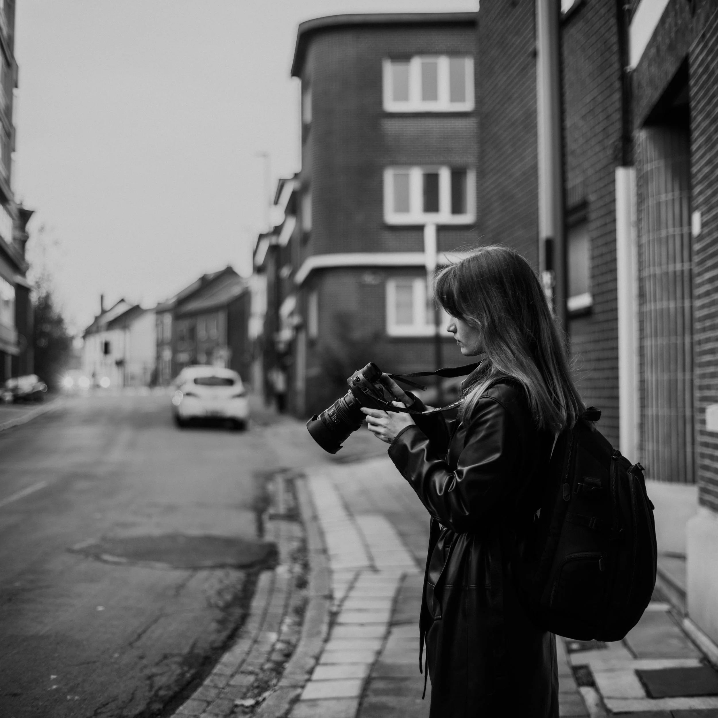 A young woman with long hair and wearing a leather jacket stands on a city sidewalk, looking at her phone. The streets are lined with brick and wooden buildings, and a car is visible in the background.