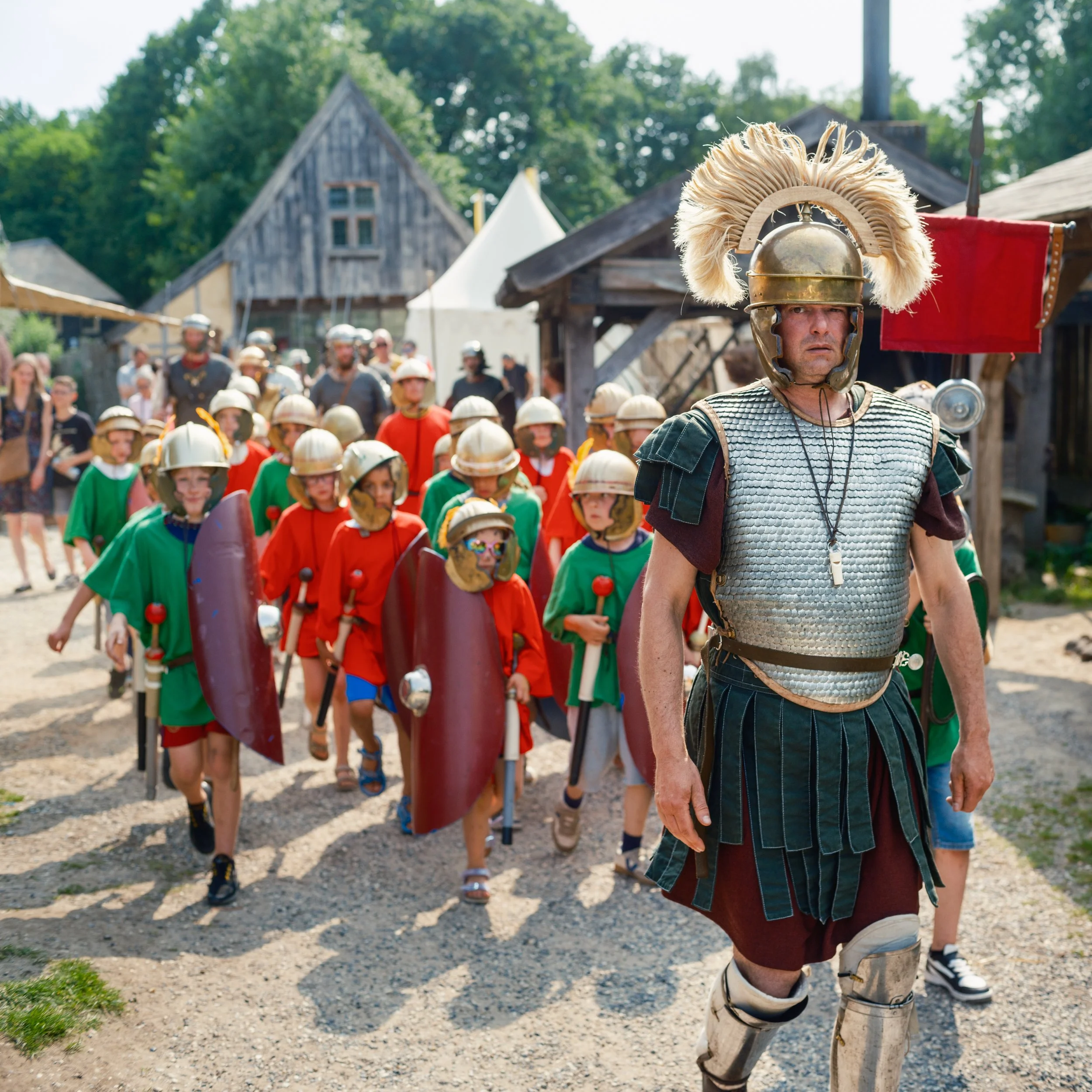 A parade or historical reenactment with people dressed as Roman soldiers, led by a man in Roman armor with a large feathered helmet, walking on a dirt path in a rural setting with wooden buildings and trees in the background.