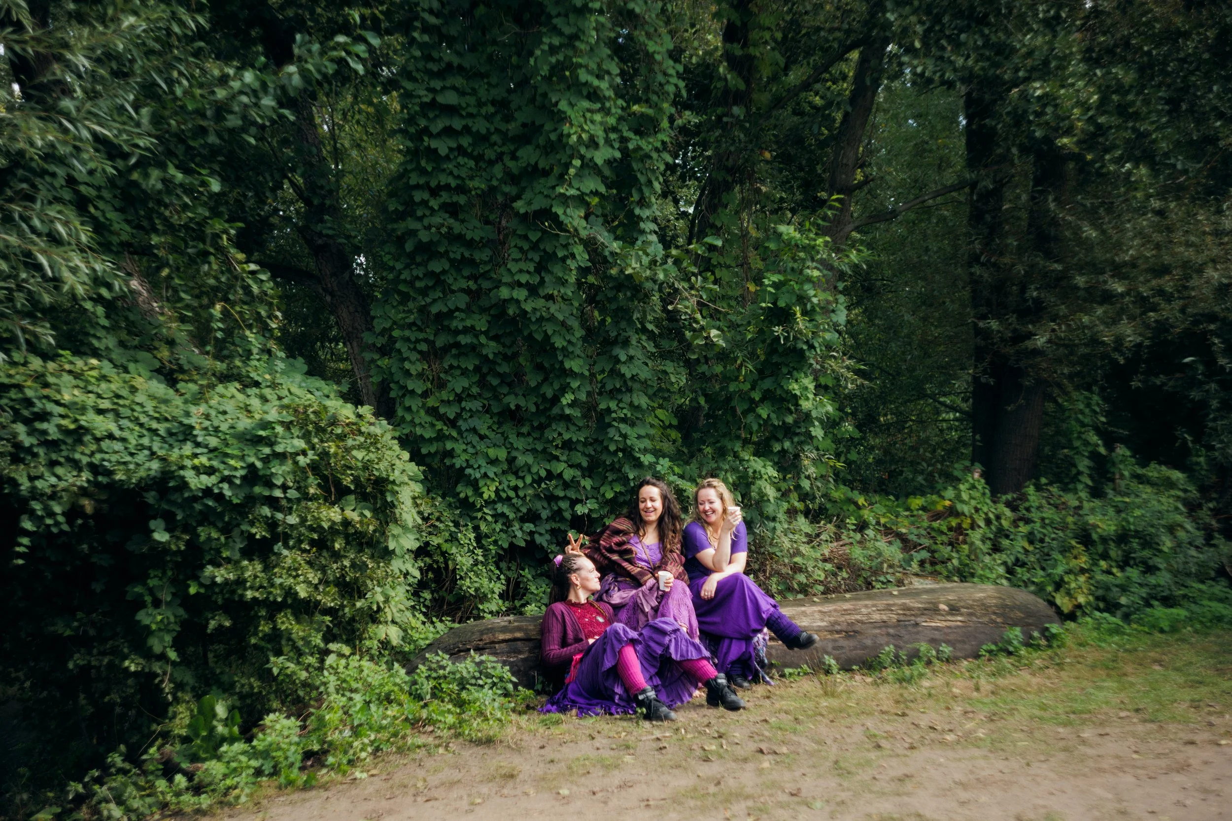Four women dressed in purple sitting on a log in a lush green forest, smiling and enjoying each other's company.