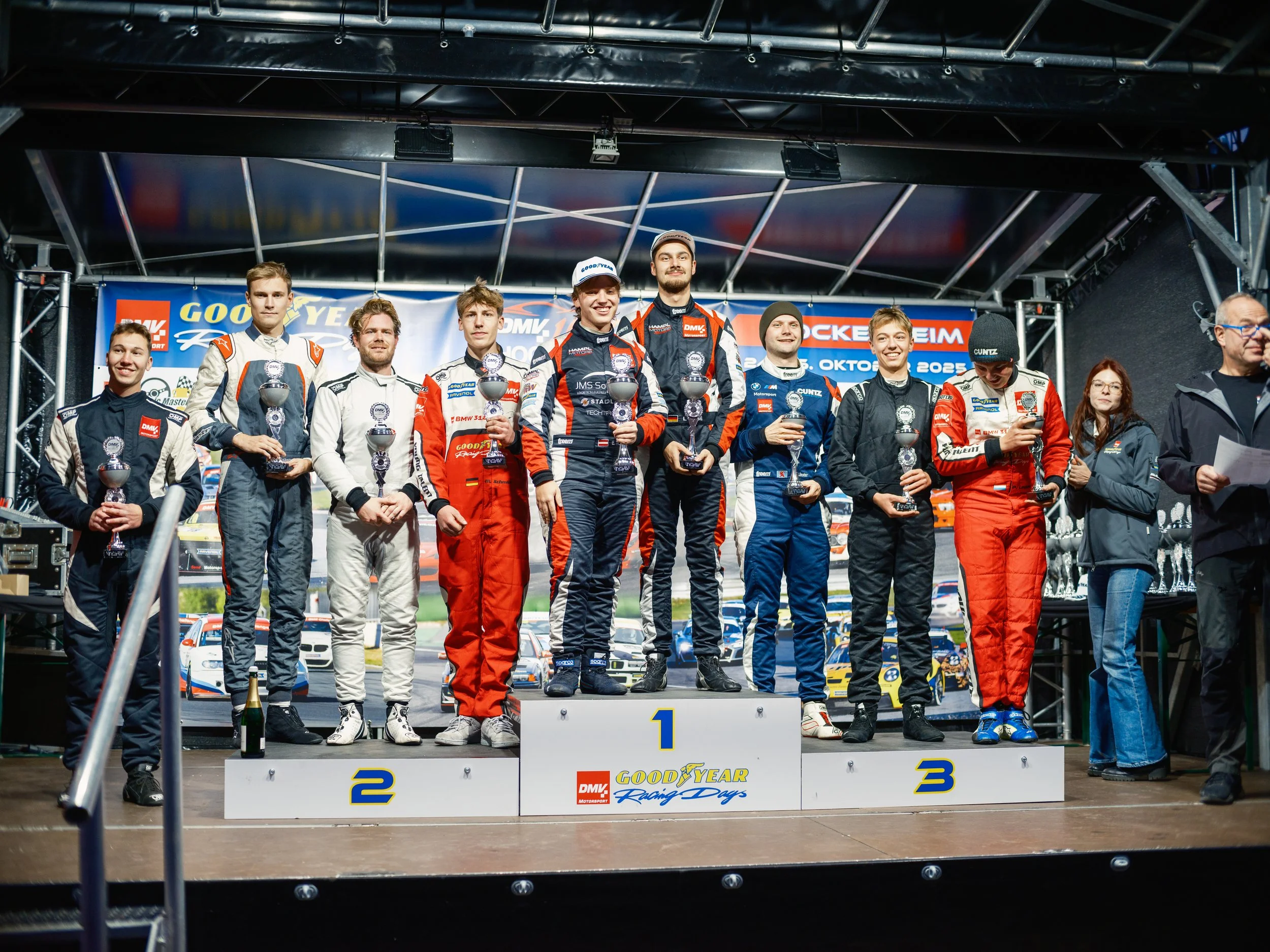 Race car drivers standing on a winners' podium holding trophies, celebrating their race results at an event with a background banner displaying sponsorship logos including Goodyear.
