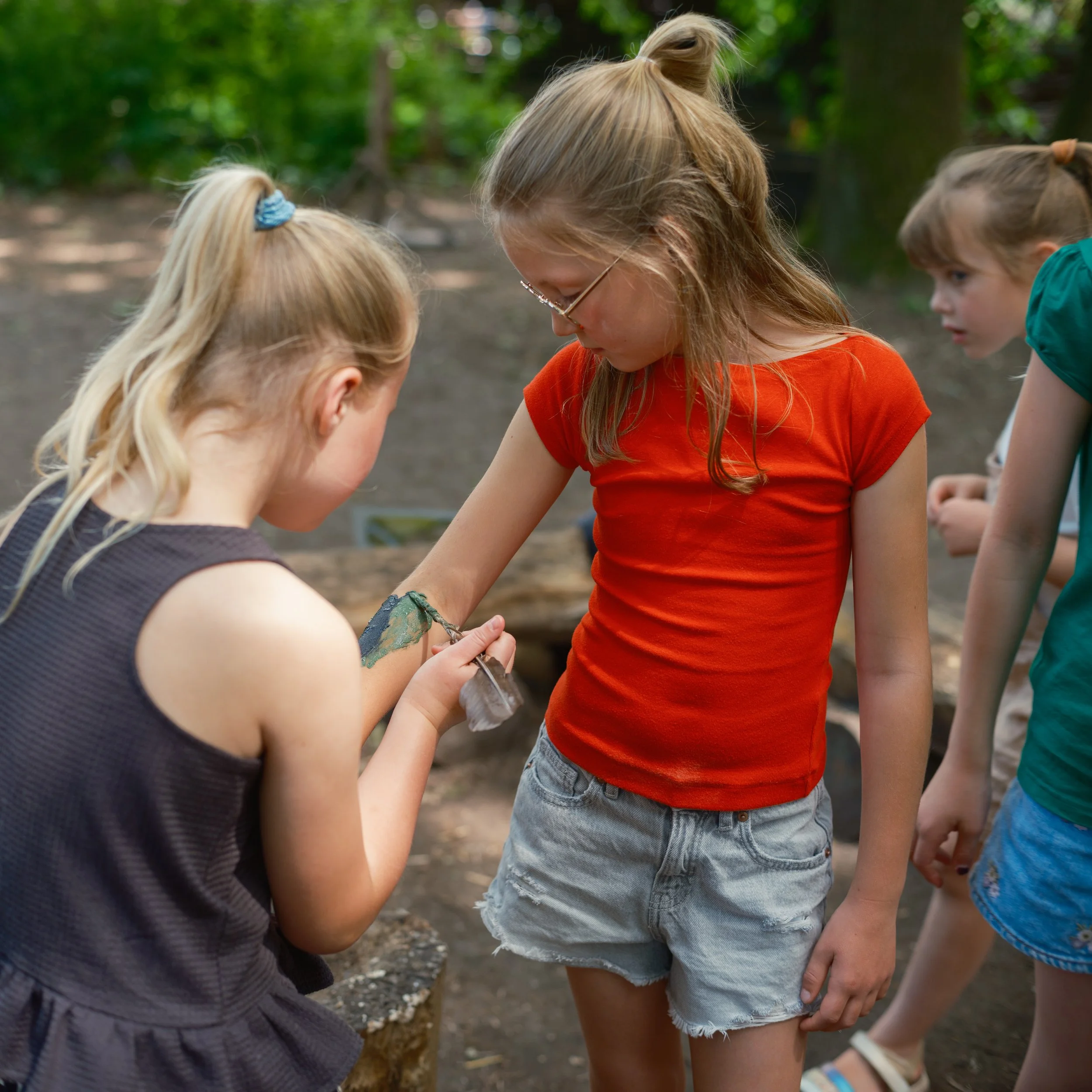Children outdoors in a park, closely examining something on the ground, surrounded by green trees.