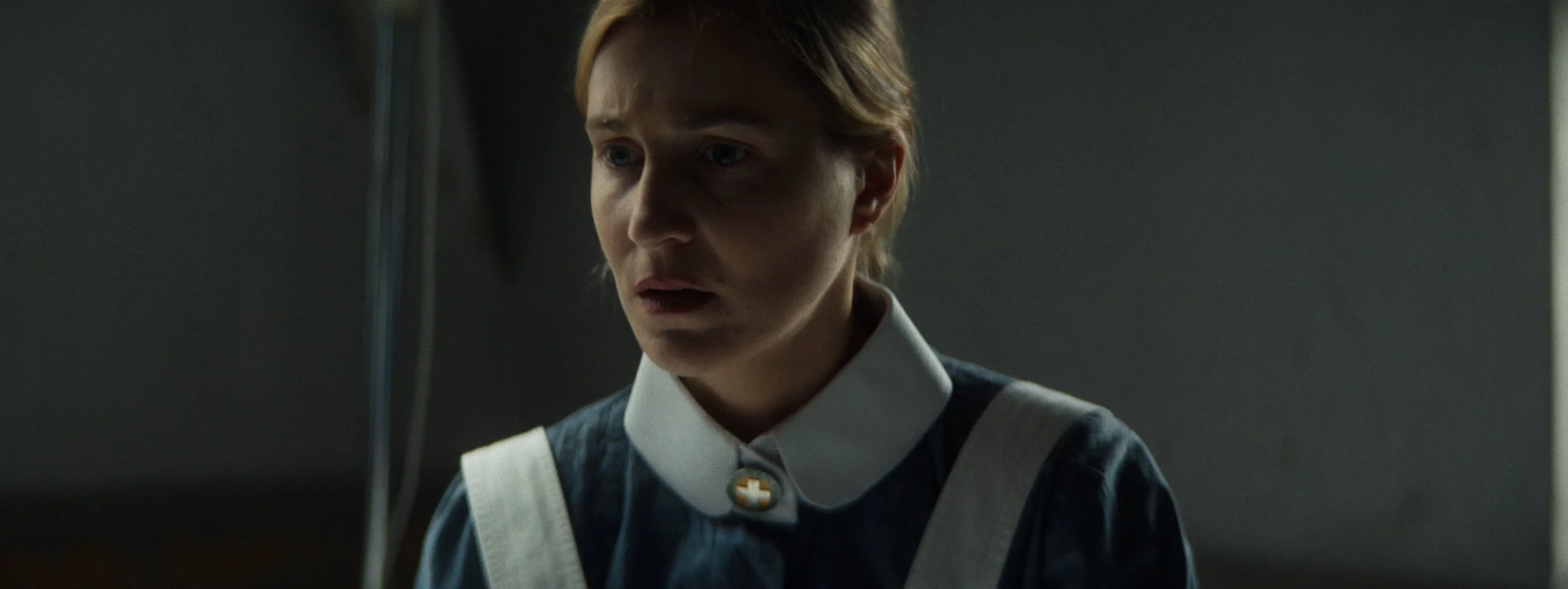 A young woman with short blonde hair, wearing a dark blue dress with a white collar and a brooch, looks down with a concerned or thoughtful expression in a dimly lit room.