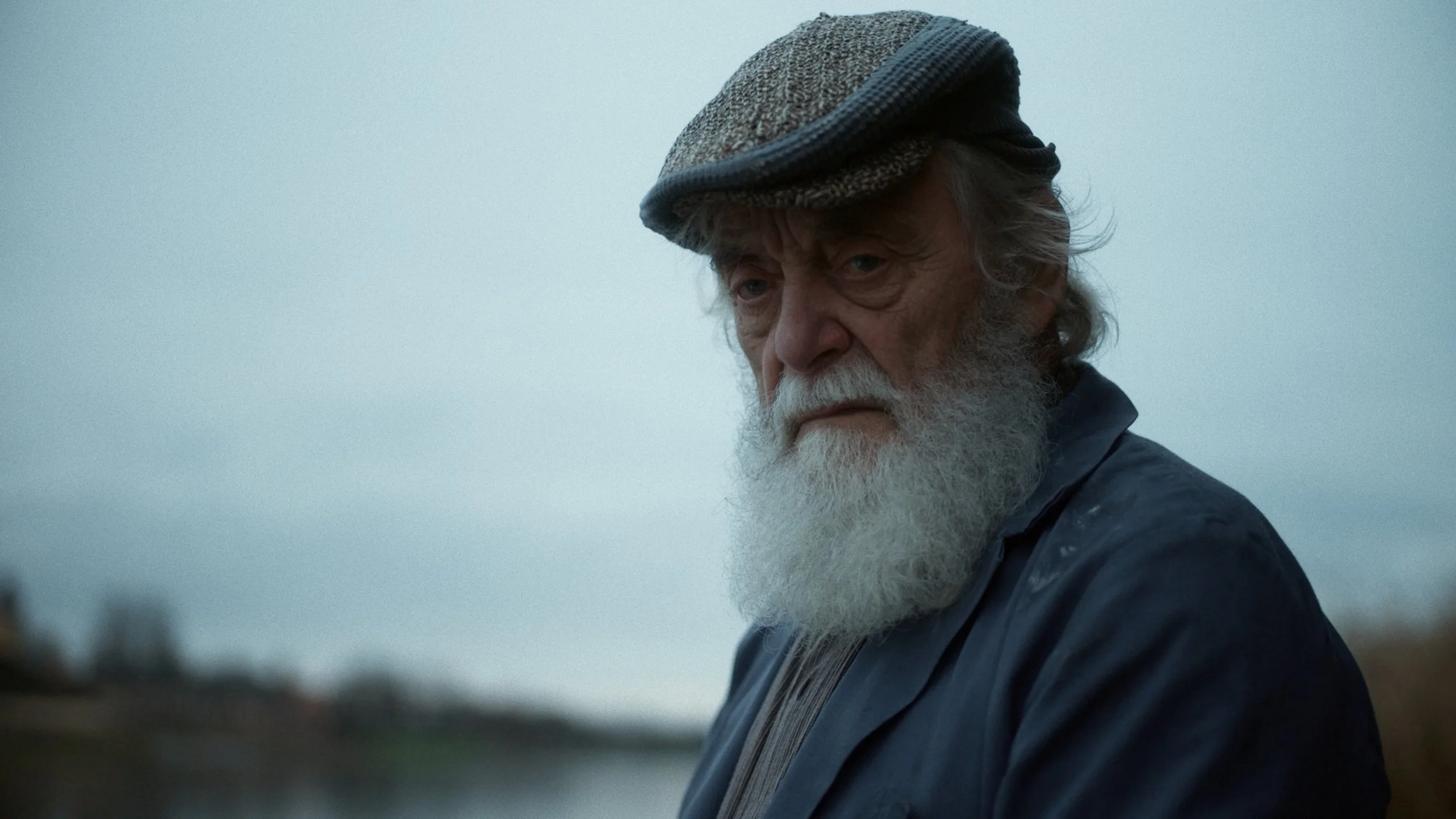 An elderly man with a white beard wearing a flat cap and a dark jacket, looking intensely with a serious expression against a cloudy sky background.