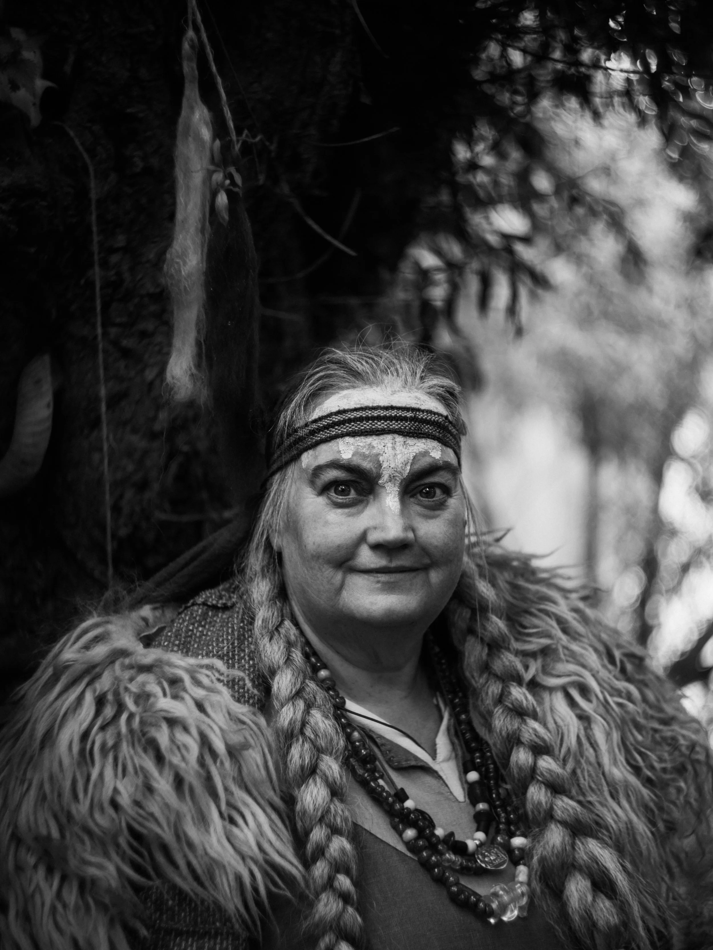 A person with long braided hair and face paint, wearing a fur coat and multiple beaded necklaces, standing outdoors with blurred trees in the background.