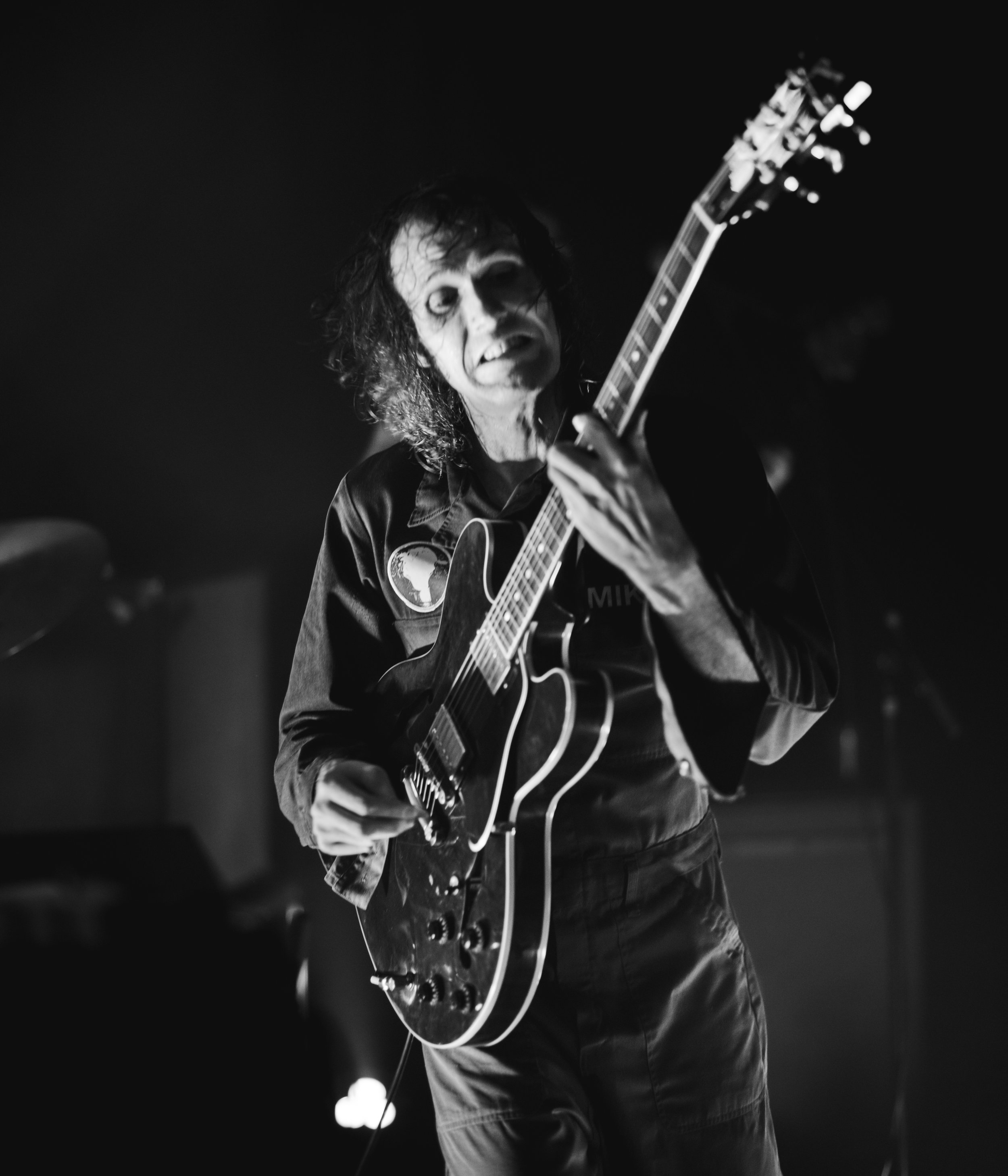 Black and white photo of a man with curly hair and sunglasses playing an electric guitar on stage. The background is dark with a few blurred lights.