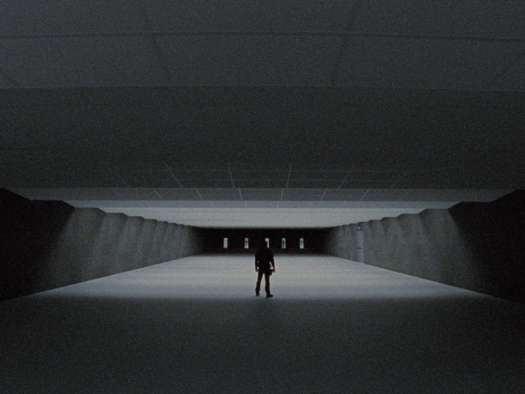 A person standing alone in a large, empty, dark tunnel with a white floor and small openings at the end, illuminated by light from behind.