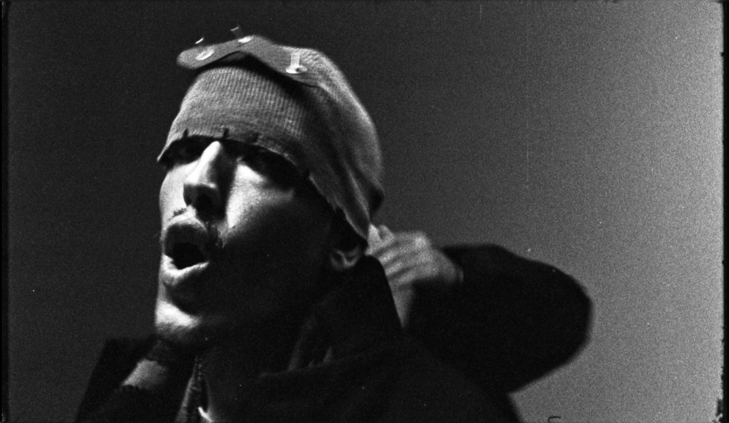 A black and white photograph of a man wearing a knit beanie and sunglasses, with a serious expression, against a dark background.