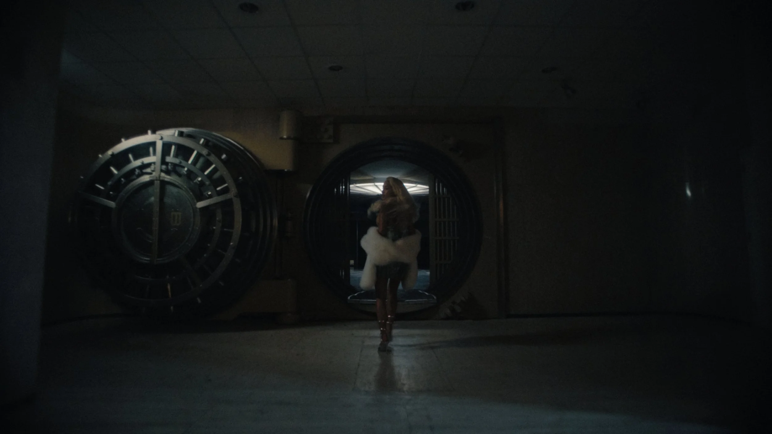 A woman standing in front of large vault doors in a dimly lit room, with her back to the camera, wearing a jacket with a hood and boots.
