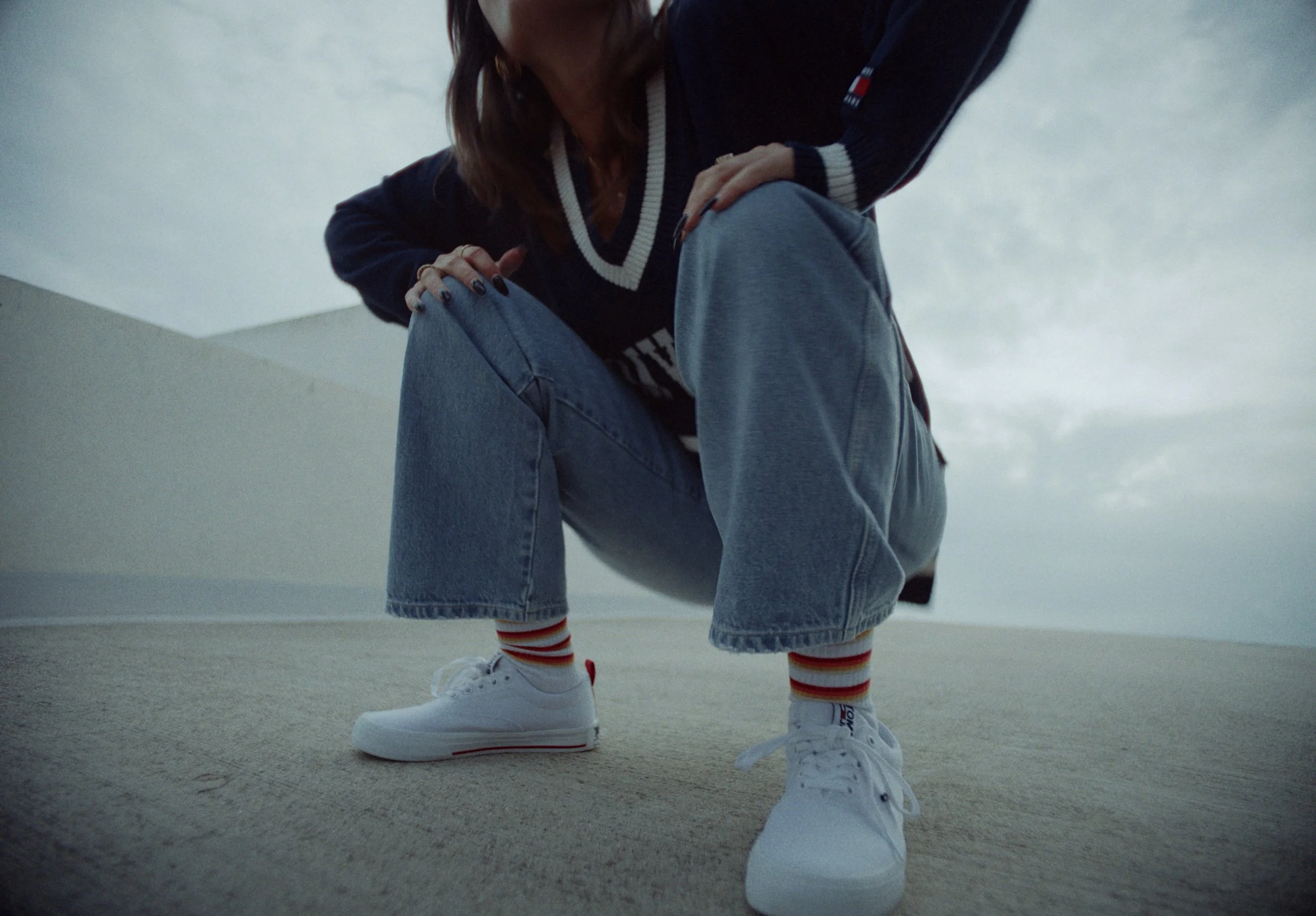 Person wearing white sneakers with red and black striped socks, loose jeans, a black jacket with red, white, and blue accents, crouching or sitting on a beige carpeted floor in a room with white walls and ceiling.