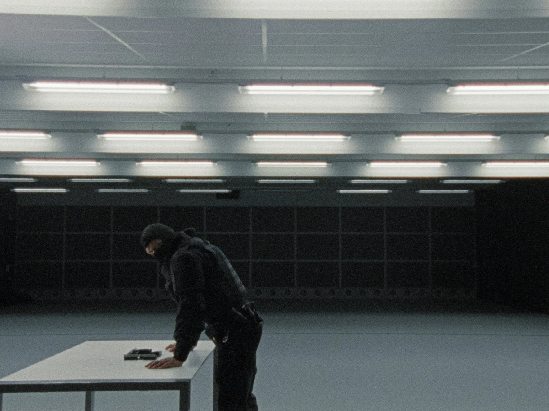A person wearing a black hoodie and face mask, bending over a table in an underground space, with a gray floor and ceiling, illuminated by ceiling lights.