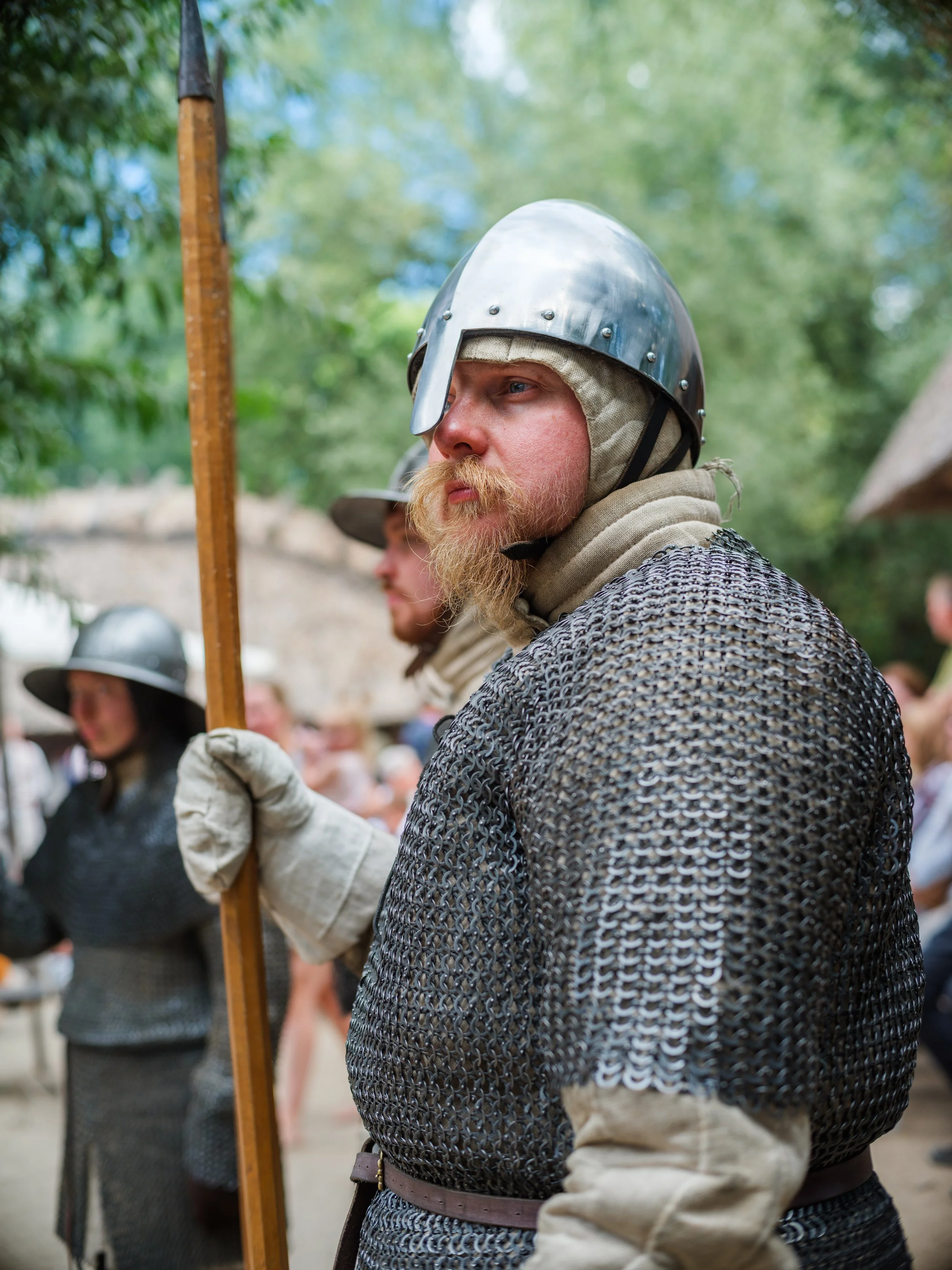 Man dressed in medieval chainmail armor, wearing a metal helmet with a leather strap, holding a wooden spear, with other people dressed similarly, outdoors with trees and a blurred background.