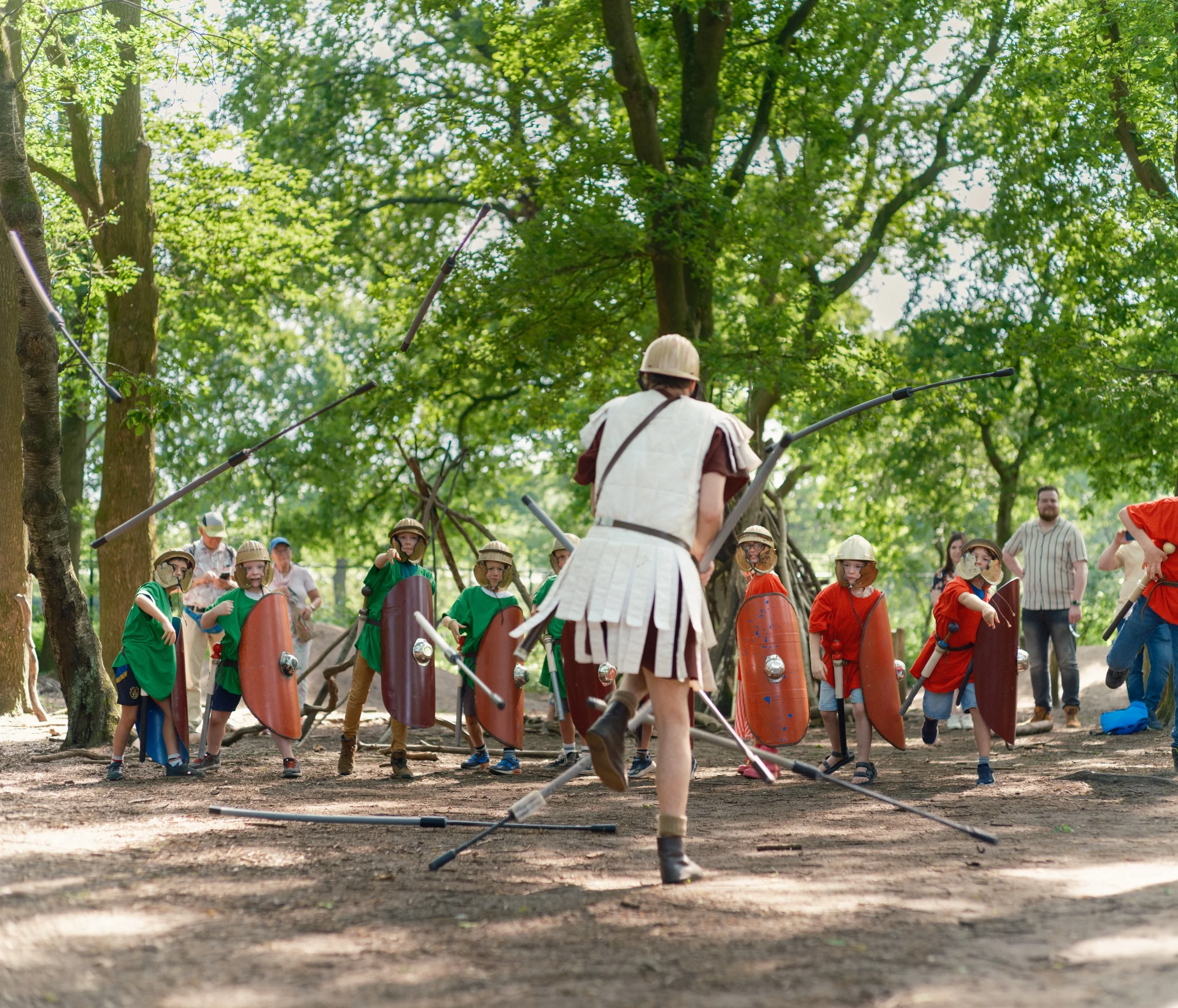 Group of children and adults participating in a medieval reenactment or educational event in a park with tall trees, some children wearing helmets and holding shields, and a woman dressed in historical costume leading the activity.
