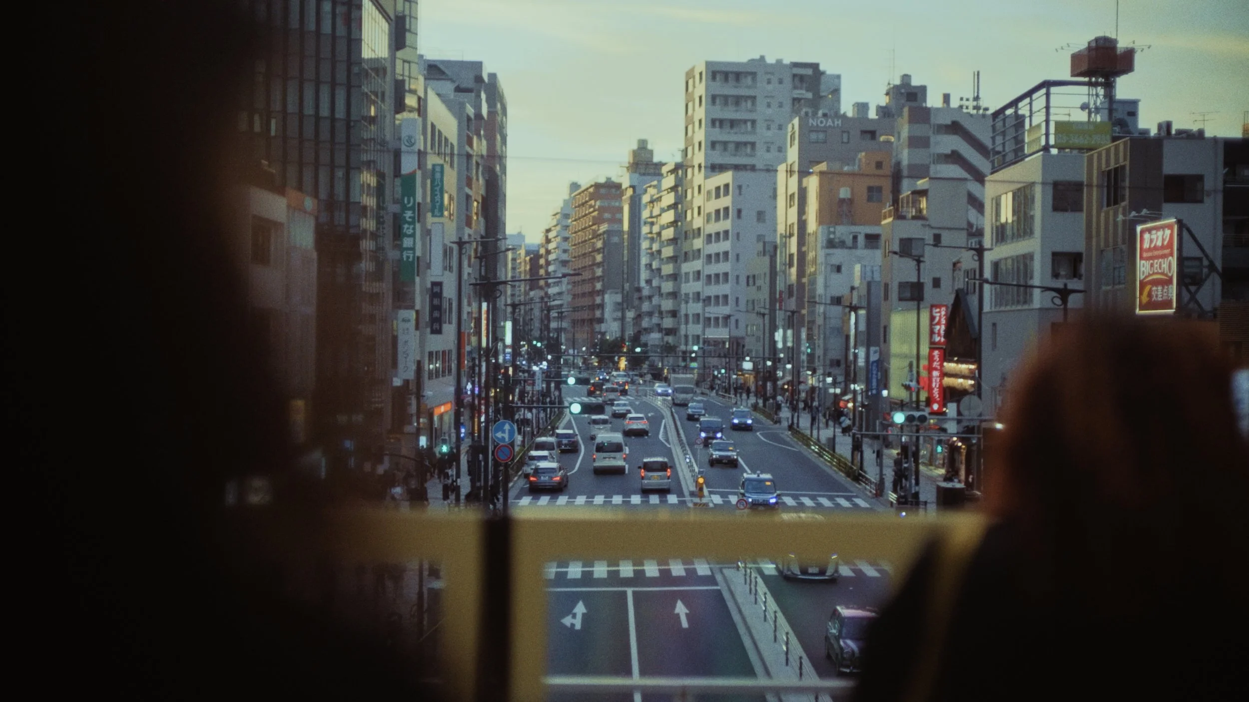 A city street viewed from a window, with tall buildings on either side of the road, multiple cars driving, and various signs with Japanese characters. The scene is during early evening or late afternoon with a soft sky.