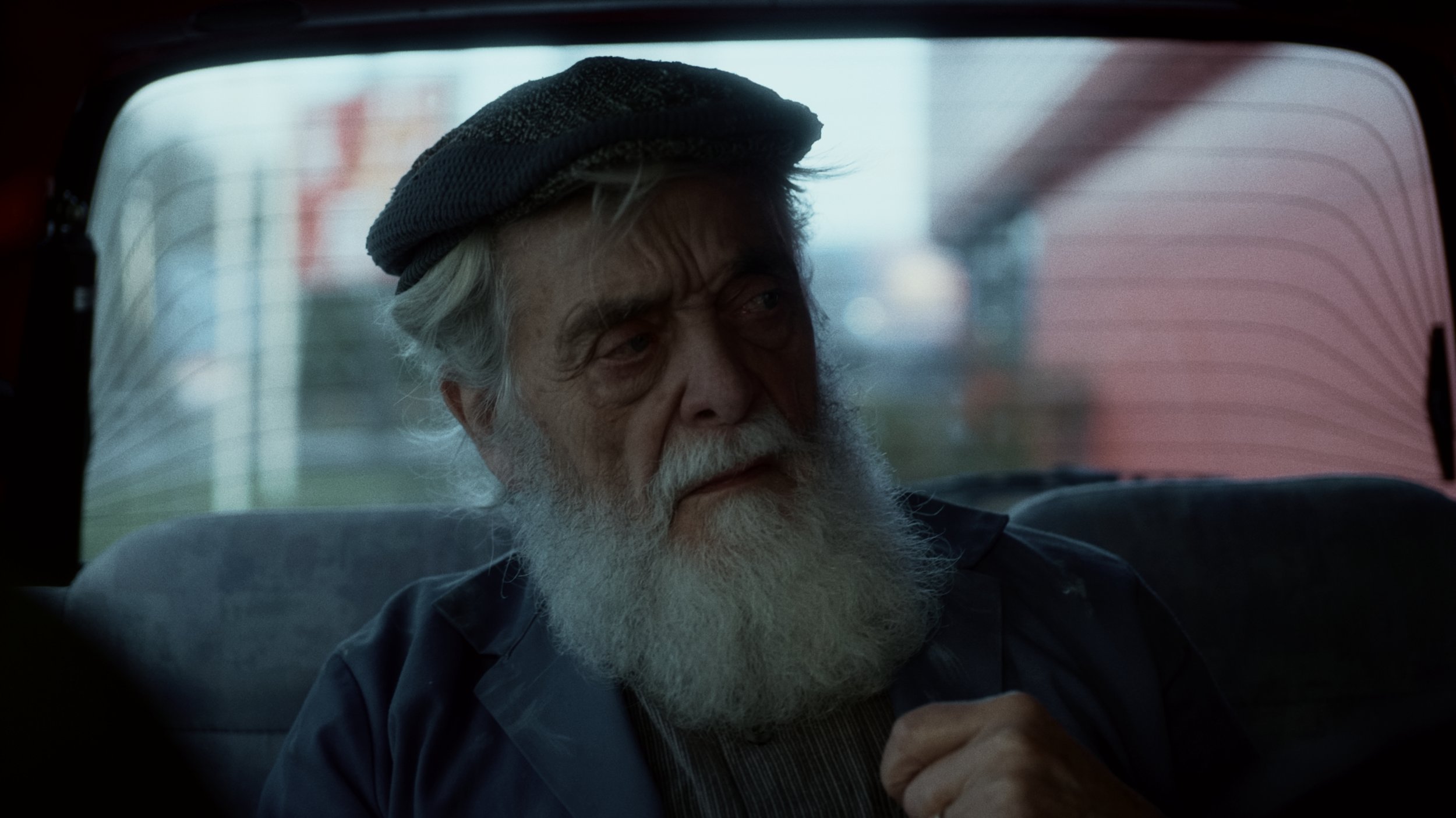 An elderly man with a white beard and grey hair wearing a grey flat cap, sitting in the back seat of a vehicle, looking out the window with a serious expression.