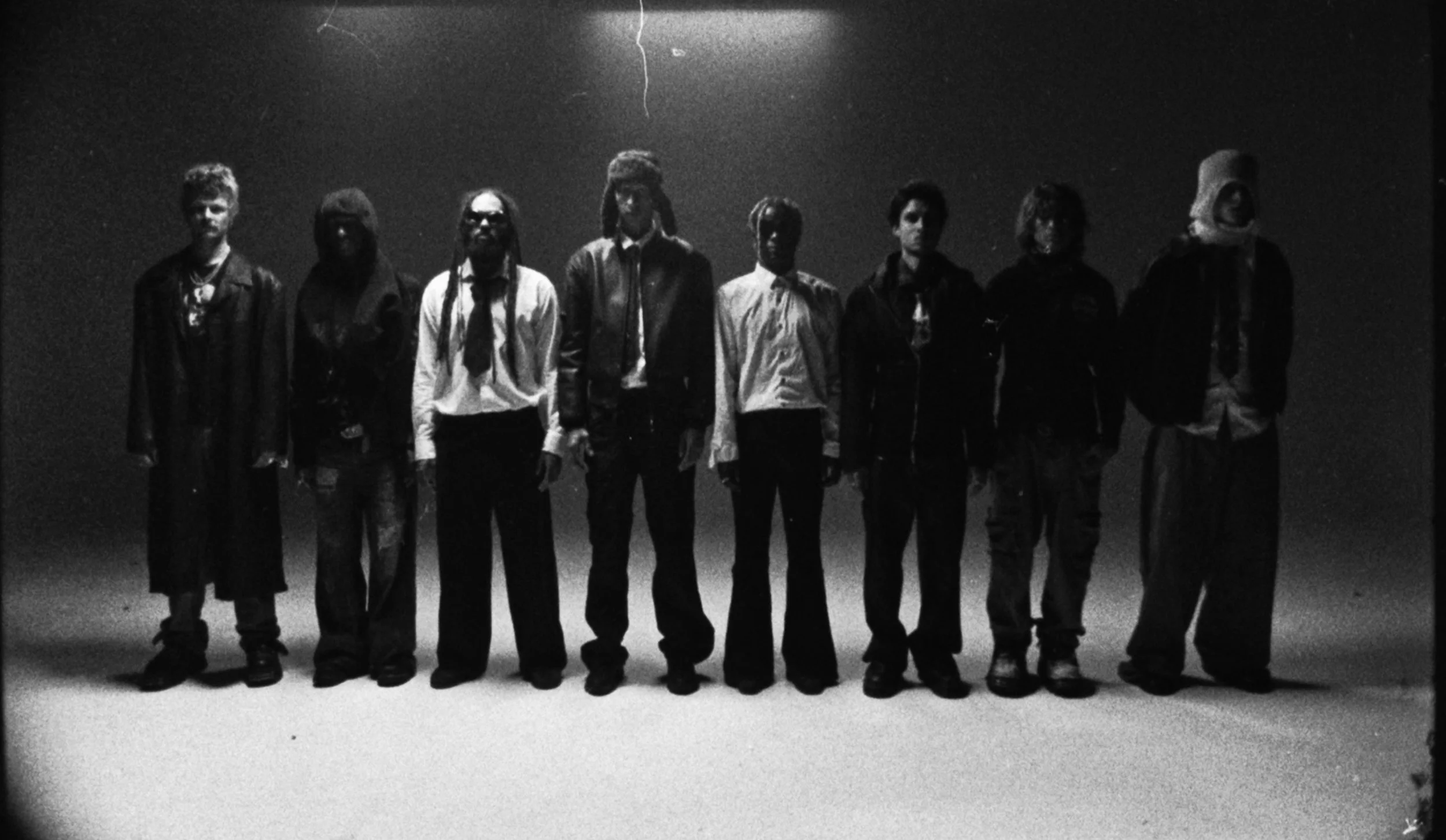 Black and white photo of eight people standing together, wearing casual and layered clothing, in a dimly lit setting with a plain background.