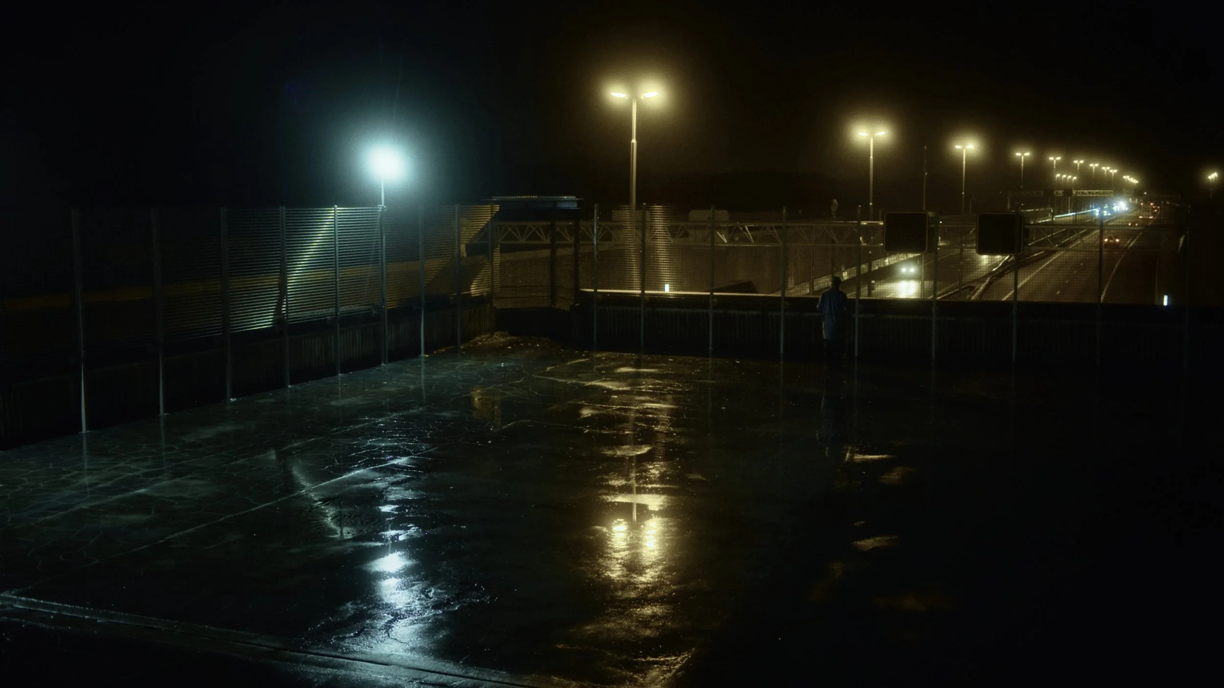A view of a bridge at night with bright streetlights reflecting off wet pavement, and a person walking near the railing.